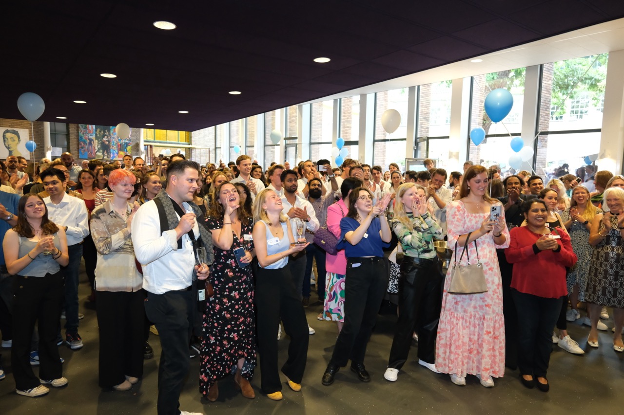 A large diverse group at an indoor event applauding and smiling with blue and white balloons in the background.