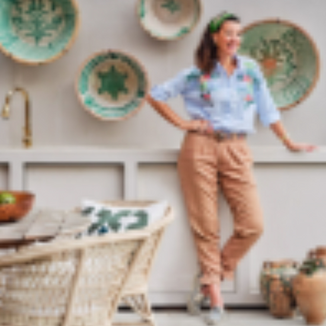 Mabel Muijres standing in a decorated room, wearing a blue embroidered shirt and tan pants, with woven bowls on the wall.