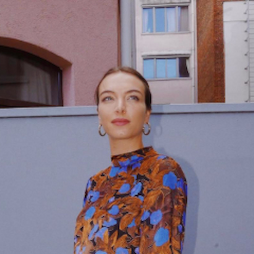 Julie Willems wearing hoop earrings and a blue and orange floral blouse, standing outdoors in front of a building.