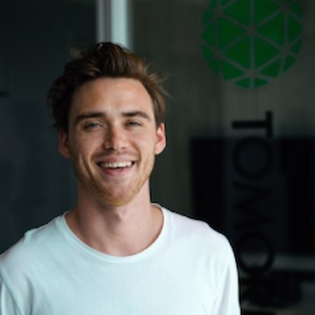 Sebastian Duyvendak smiling in a white t-shirt, standing indoors in front of a glass wall with a green logo.