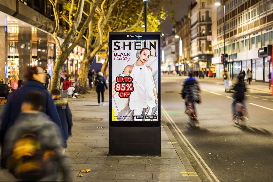 Bus stop digital advertisement for SHEIN Black Friday sale offering up to 85% off, on a city street at night with pedestrians and cyclists.