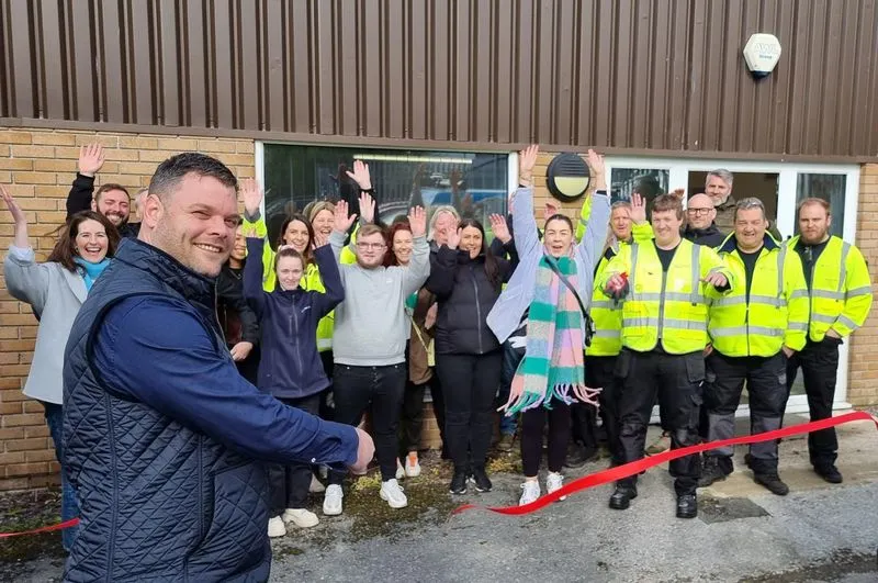 Group of people with some in yellow safety jackets cheering and waving behind a man cutting a red ribbon in a ribbon-cutting ceremony outside a building.