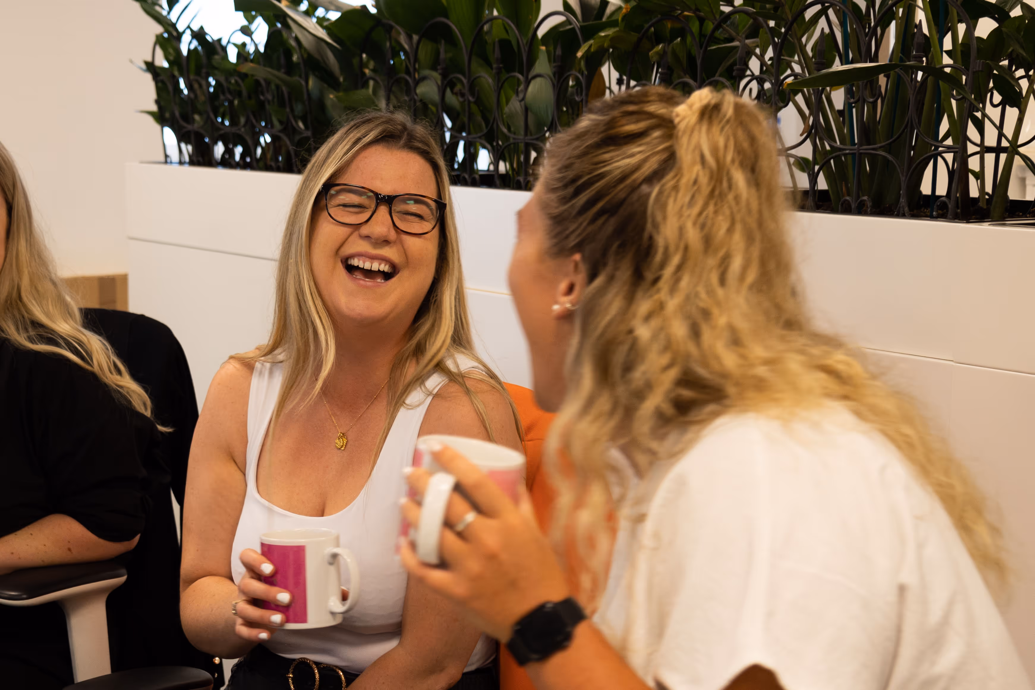 Two women laughing and holding coffee mugs in a casual indoor setting with plants in the background.