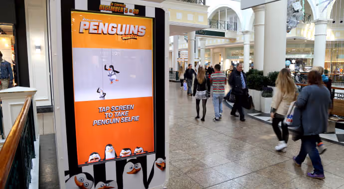 A Penguins movie ad on a digital screen in a busy shopping mall