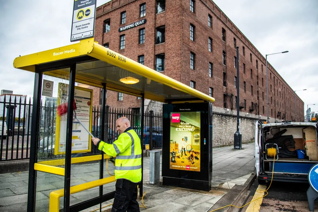 A male Bauer Media Outdoor construction worker cleaning a yellow framed bus shelter in front of a red brick building