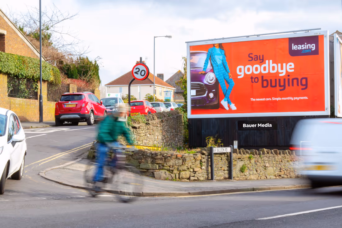 Bauer Media Outdoor digital advertising billboard on a residential roadside displays a leasing.com ad with the message "Say goodbye to buying," as cars and a cyclist pass by.
