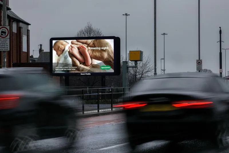 Bauer Media Outdoor Billboard digital advertising screen on a roadside displays a Specsavers ad featuring baby feet and the message "Amazing sight," as cars drive past.