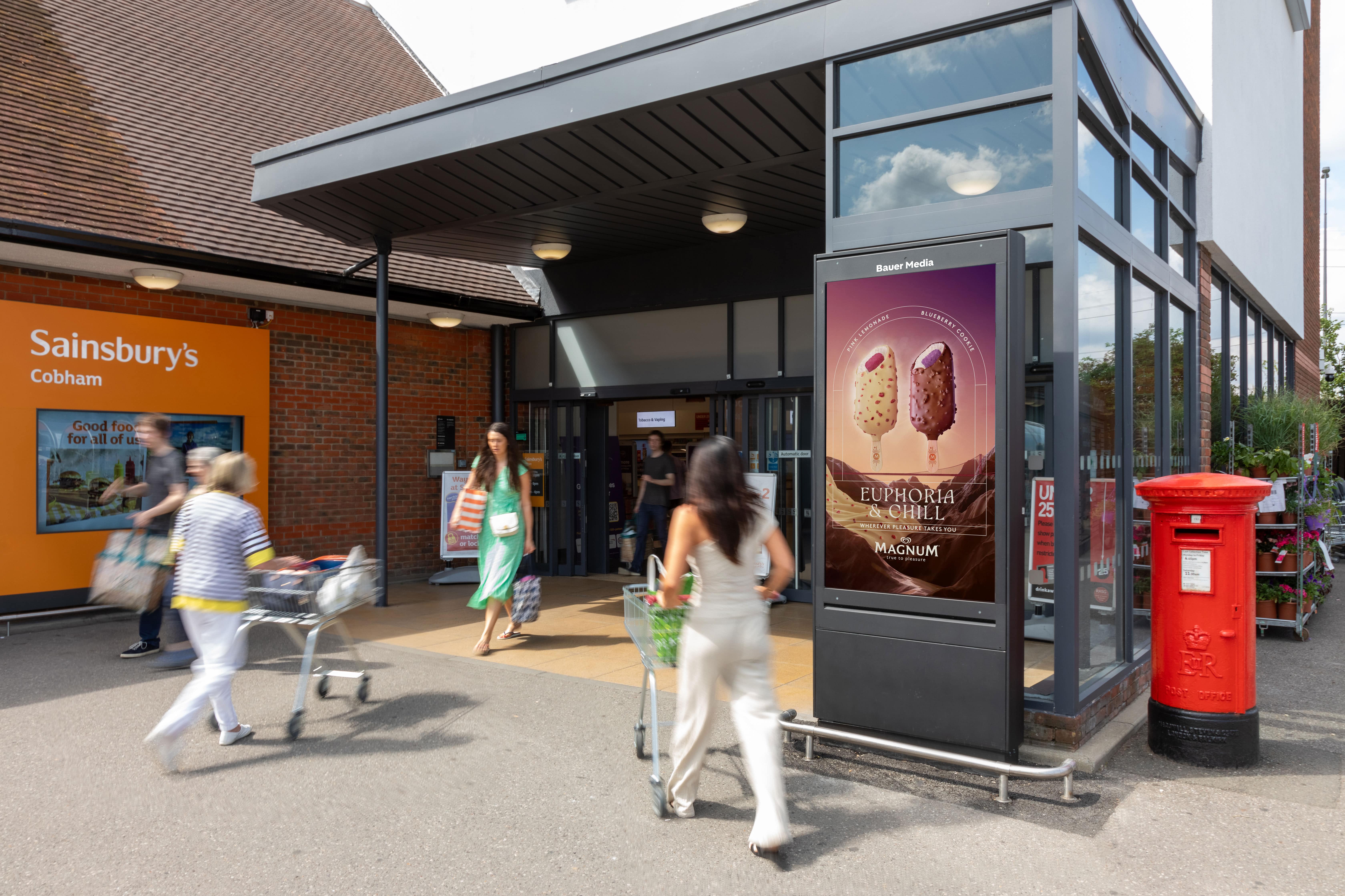 Digital advertisement outside the front of a Sainsbury's store with shoppers walking past to go into the store.