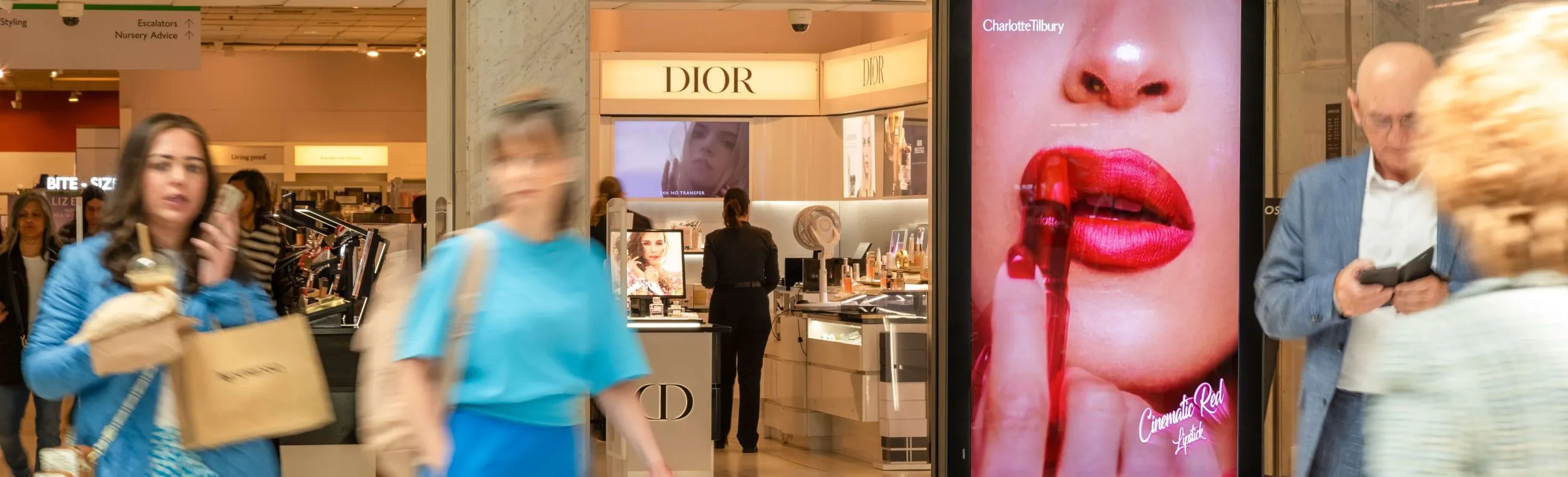 Bauer Media Outdoor Digital advertising screen in a malls displays a Charlotte Tilbury lipstick ad. Shoppers pass by, with the Dior beauty counter visible in the background.