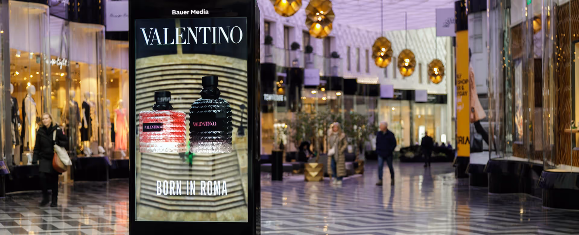 Bauer Media Outdoor Digital advertising screen in a shopping mall displays a Valentino fragrance ad. Shoppers in the background of upscale retail setting.