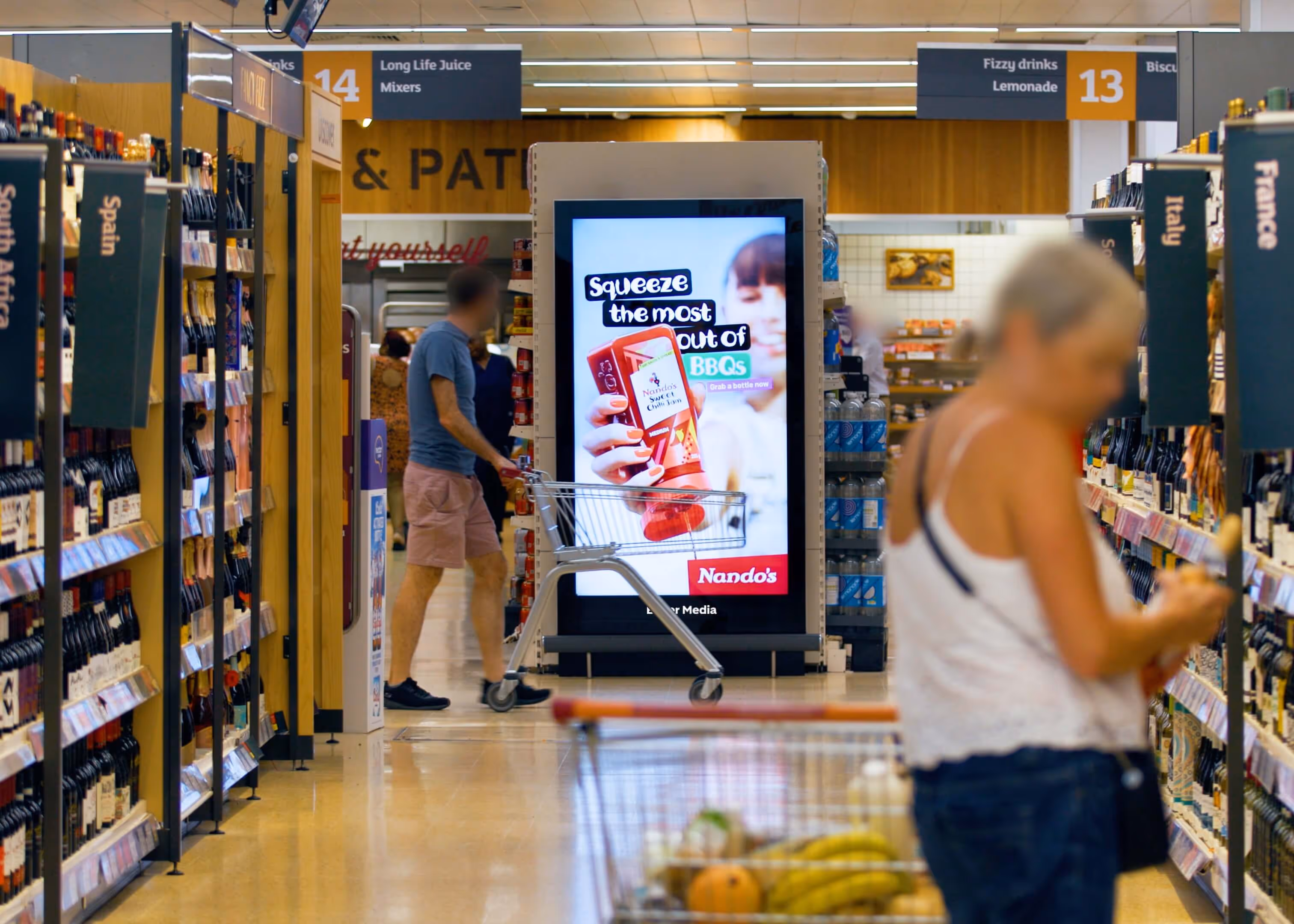 Bauer Media Outdoor digital advertising screen inside a Sainsbury's displays a Nando’s sauce ad with the message “Squeeze the most out of BBQs,” as shoppers browse the aisles.