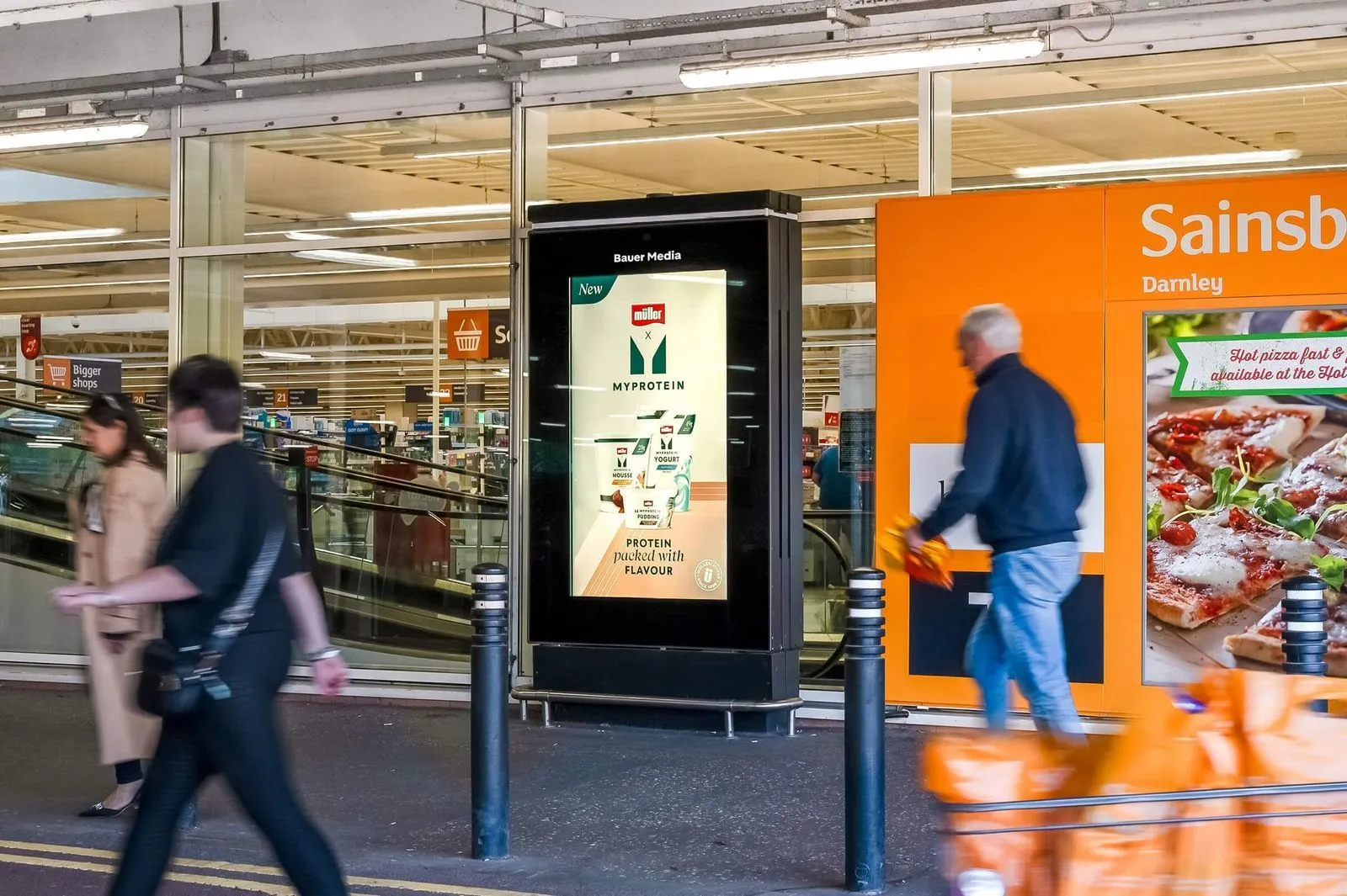 Entrance of a Sainsbury's supermarket with an advertisement for Müller x MyProtein products on a digital display and people walking by.