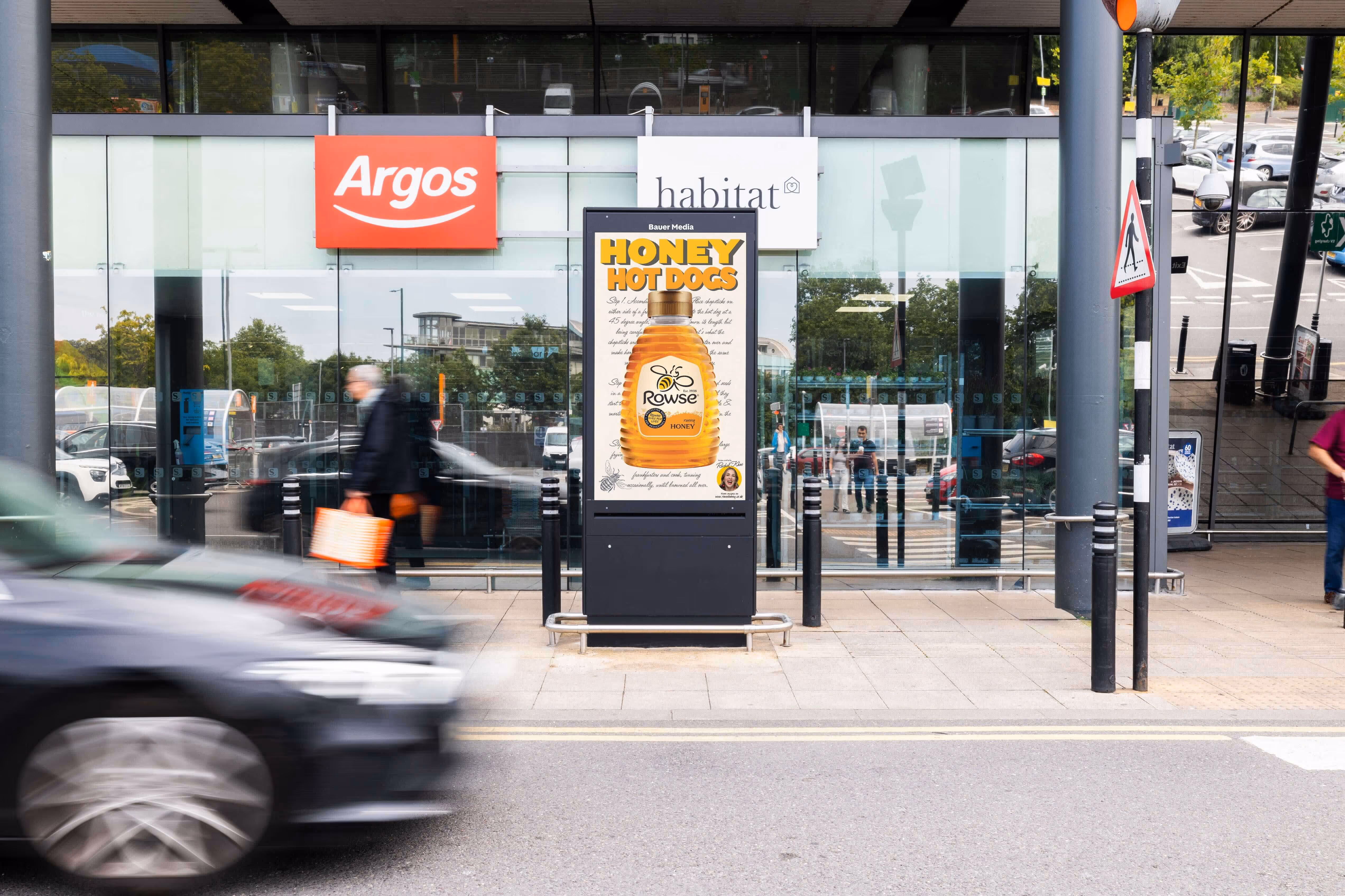 Bauer Media Outdoor digital advertising screen outside Sainsbury's displays a Rowse honey ad with the headline “Honey Hot Dogs,” as pedestrians pass by.