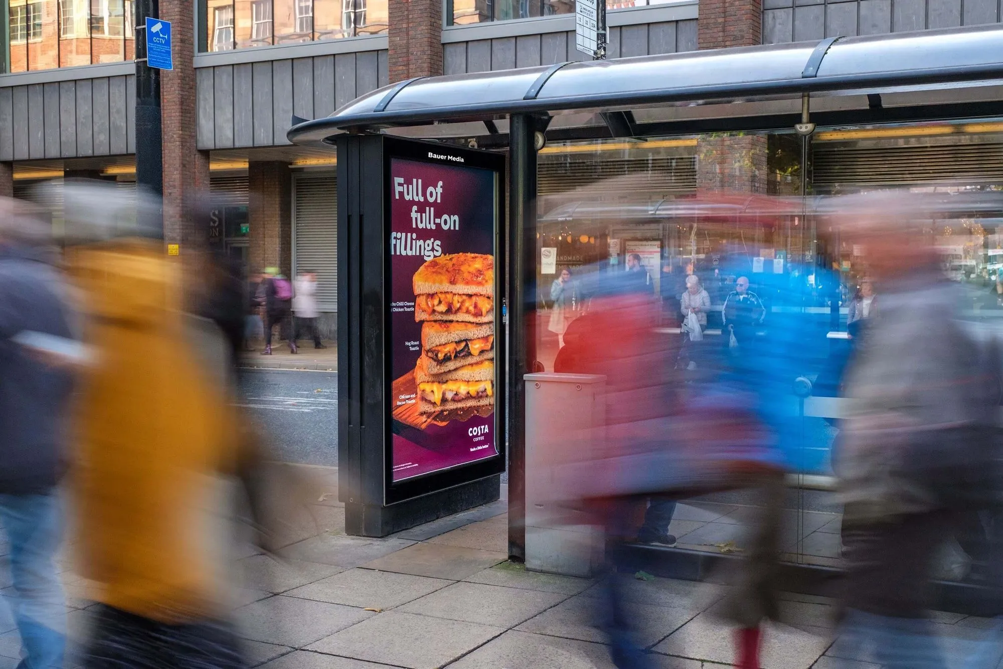 Bauer Media Outdoor digital advertising screen at a busy bus stop displays a Costa ad featuring stacked toasties with the tagline “Full of full-on fillings,” as pedestrians pass by.