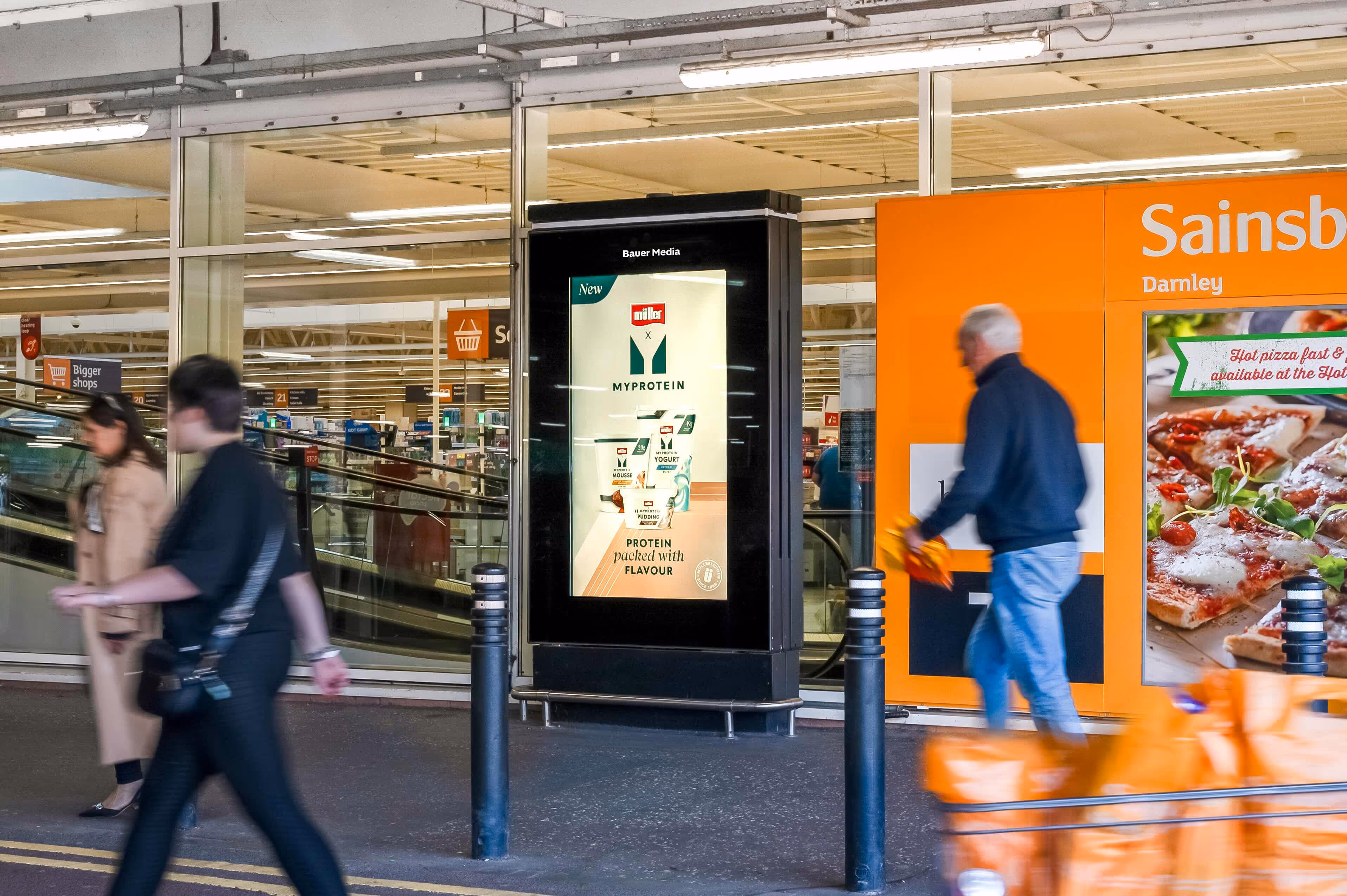 Bauer Media Outdoor digital advertising screen outside a Sainsbury’s store displays a Yeo Valley Organic campaign, as shoppers walk past with groceries.