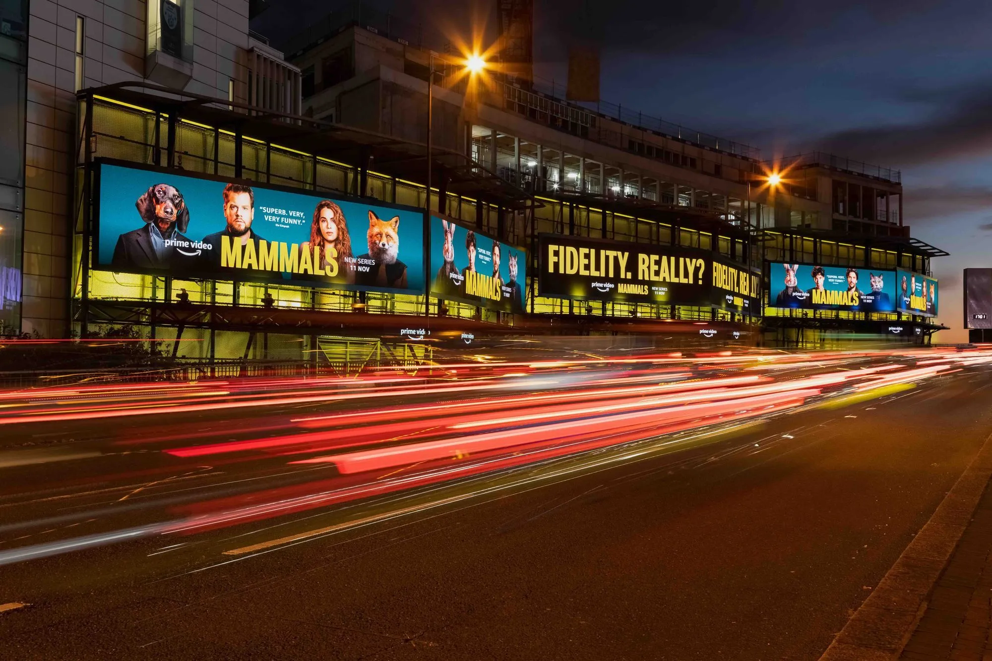 Bauer Media Outdoor digital billboards on a city building display an Amazon Prime Video campaign for the series “Mammals,” with bold text reading “FIDELITY, REALLY? RUTH?” as traffic streaks by on a busy evening road.