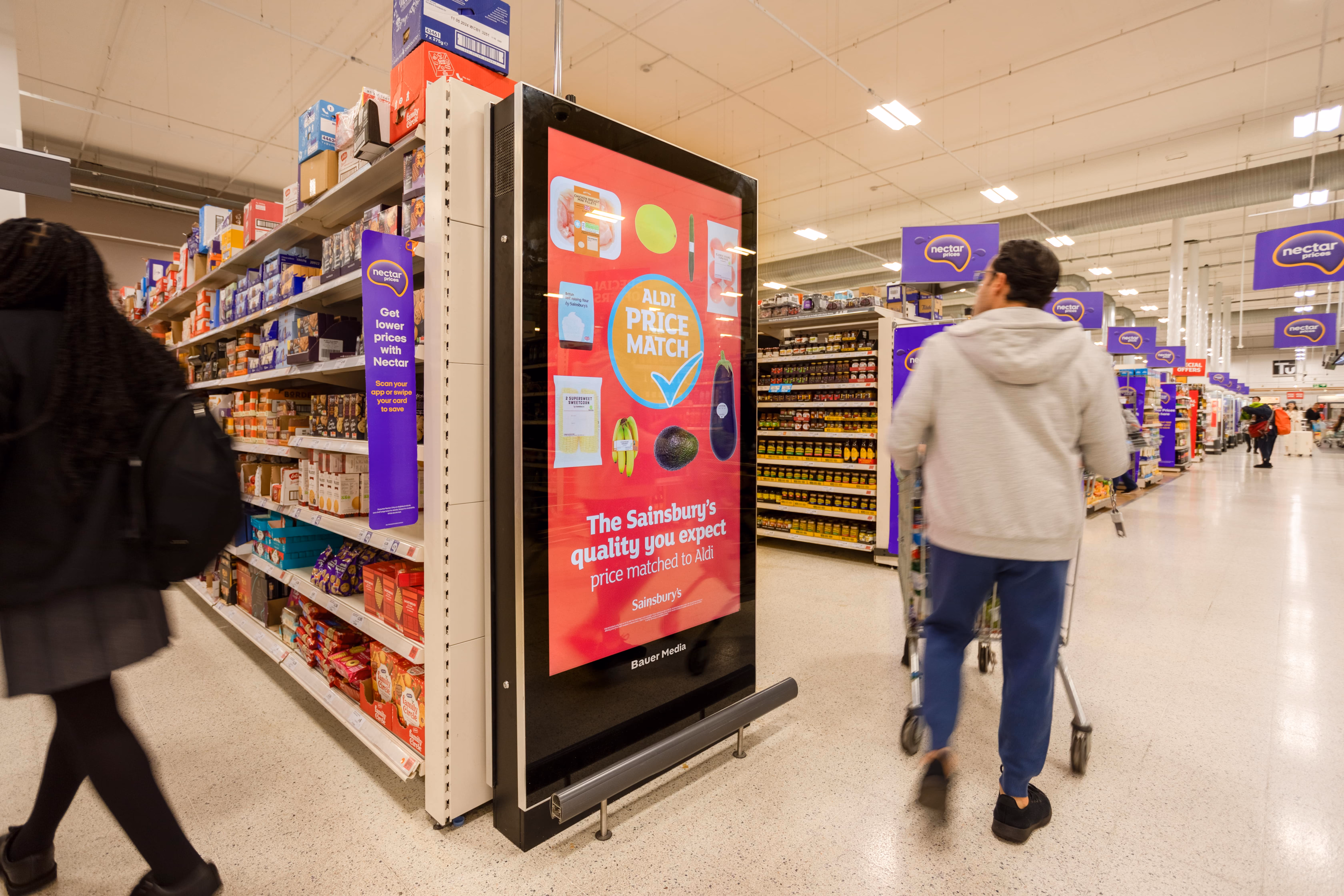 Digital sign in a grocery store aisle showing an Aldi price match advertisement with shoppers nearby.