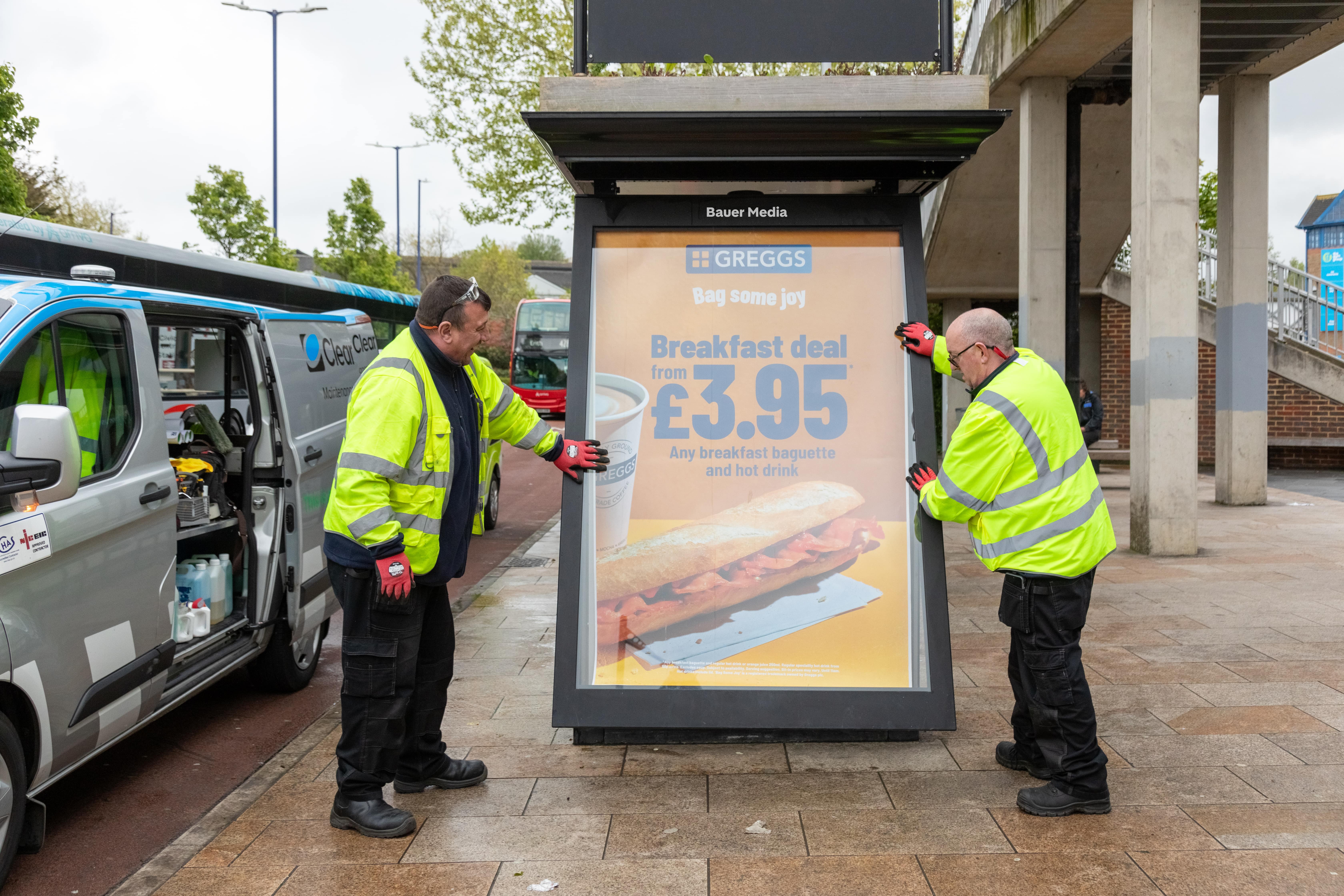 Two workers in high-visibility jackets installing a Greggs advertisement board showing a breakfast deal for £3.95 with a baguette and hot drink.