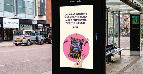 Bus stop digital advertisement showing a hand holding a book titled 'EMILY' with text stating it was meant to be seen when warmer weather arrives but is seen now.