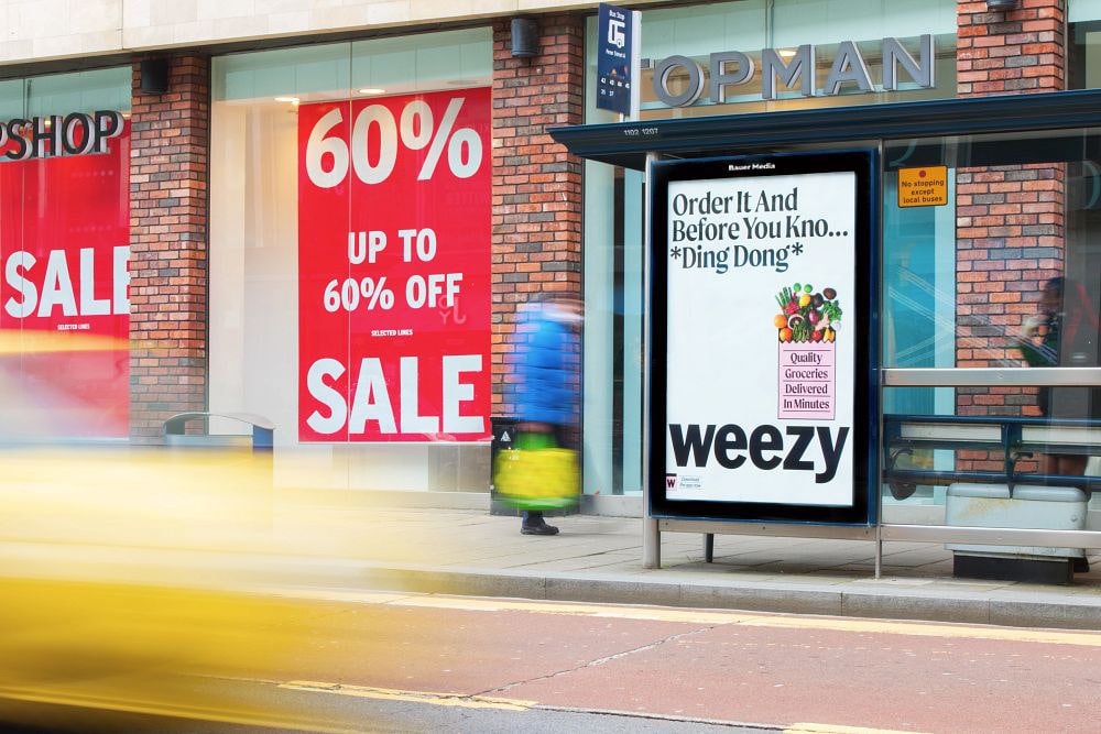 Bus stop advertisement for Weezy grocery delivery showing text 'Order It And Before You Kno... *Ding Dong* Quality Groceries Delivered In Minutes' on a white background, with a blurred yellow vehicle passing by and a large red 60% off sale sign in a shop window behind.