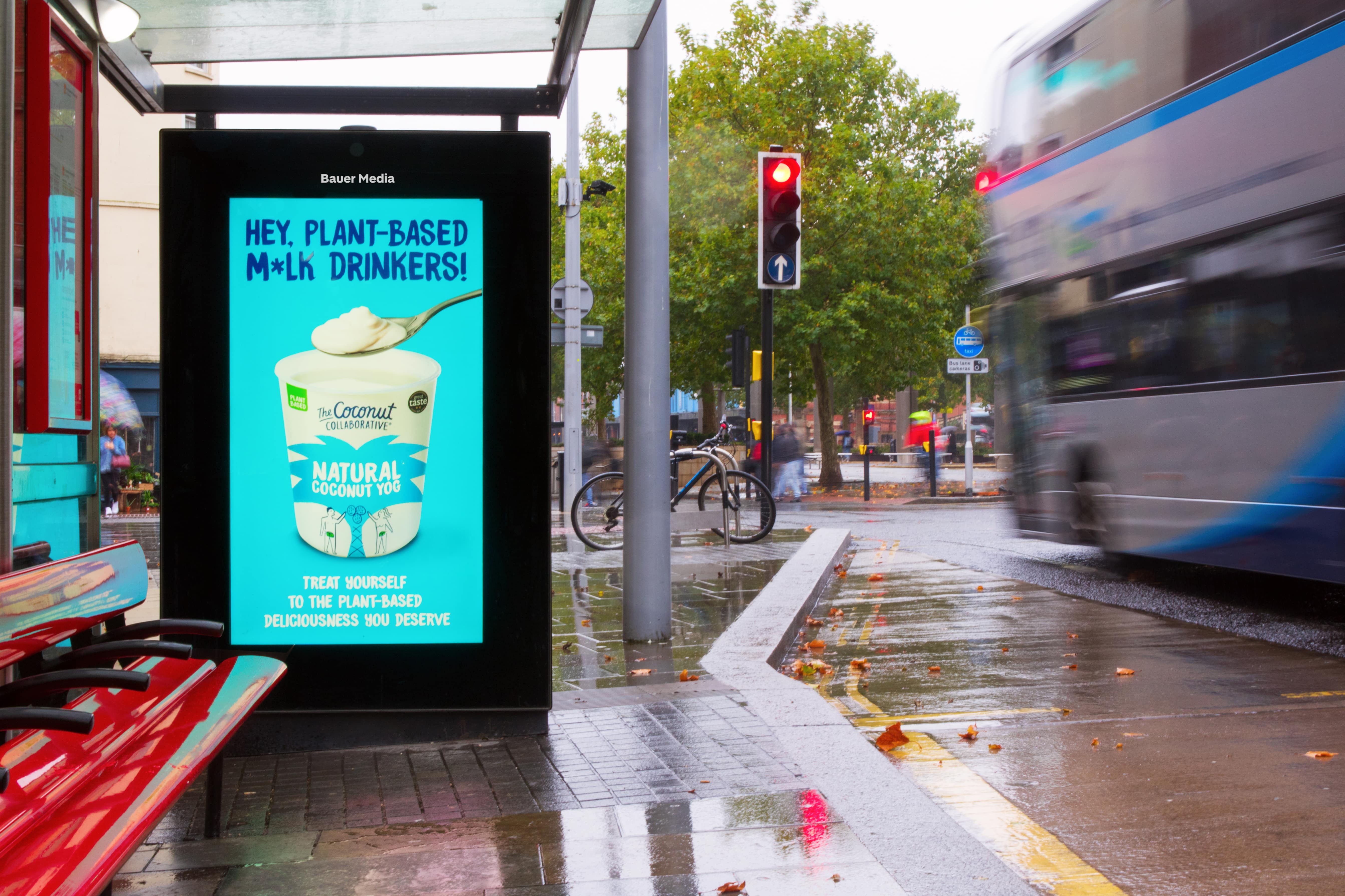 Bus stop with a digital advertisement for plant-based natural coconut yogurt on a rainy day, a bus passing by, and a red traffic light.