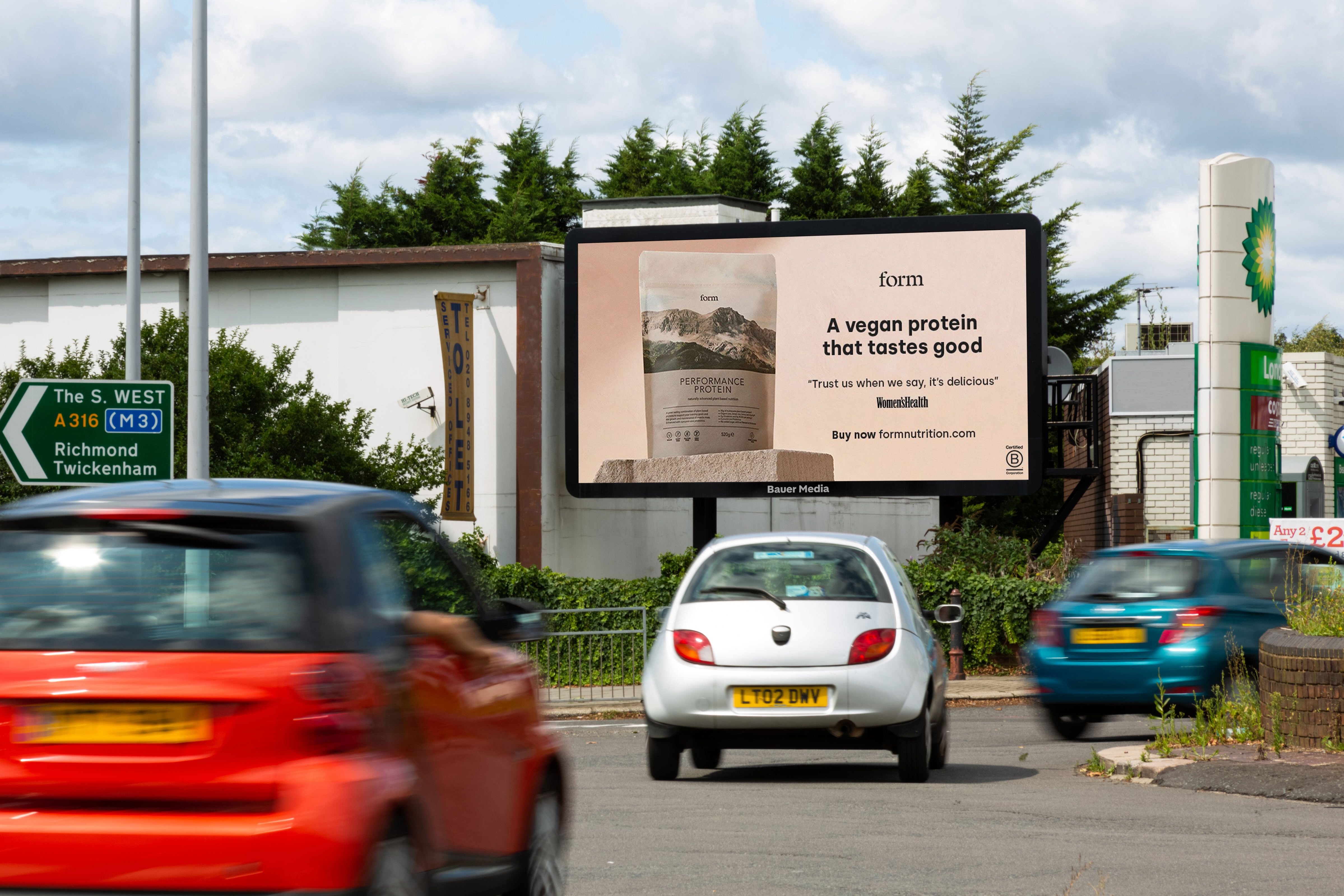 Billboard advertising Form Performance Protein, a vegan protein powder, with a quote from Women's Health and a prompt to buy at formnutrition.com, seen on a street with cars and directional signs in the background.