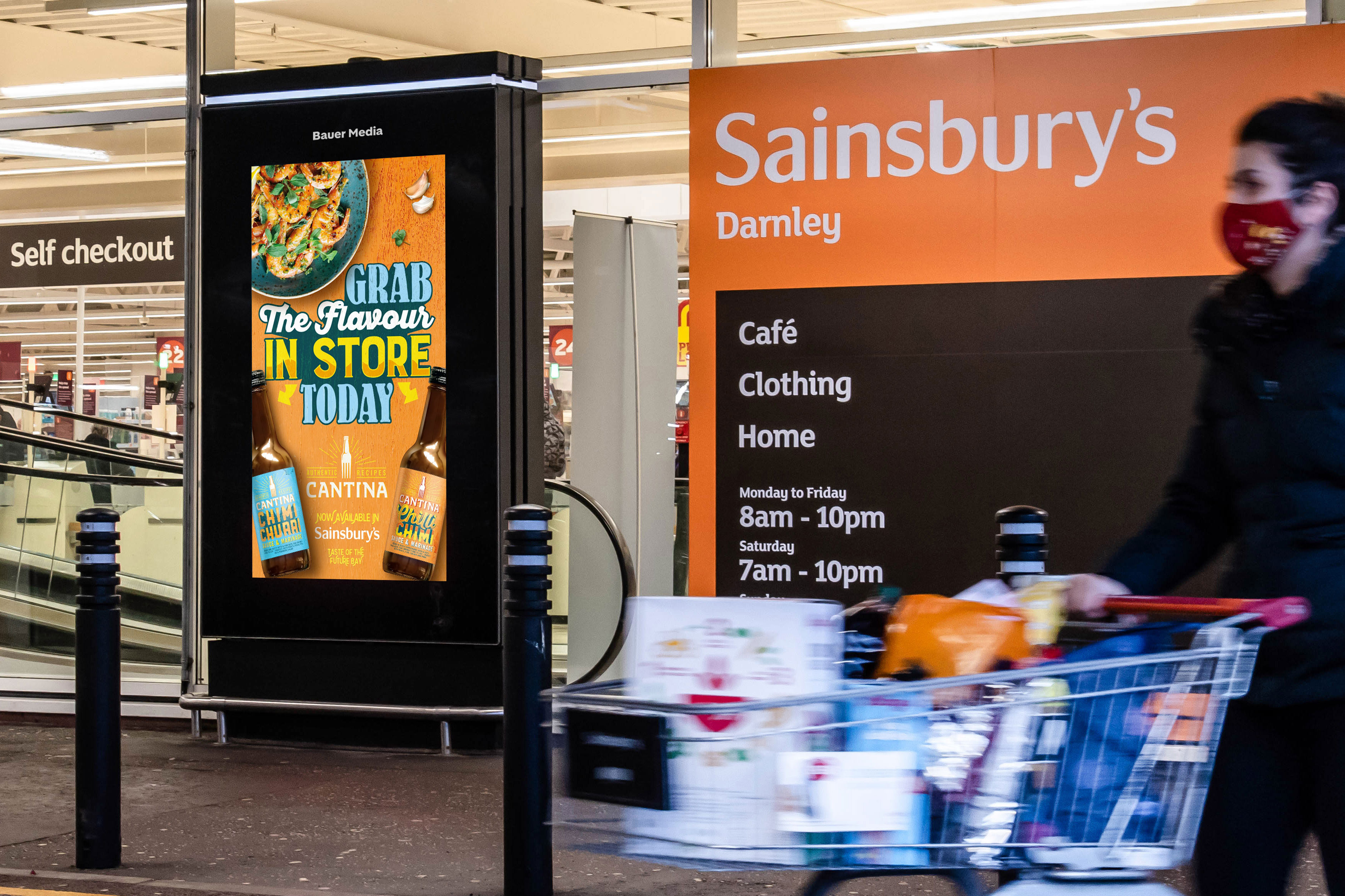 Sainsbury's store entrance showing opening hours and a digital advert promoting Cantina chimichurri sauce with a shopper pushing a cart full of groceries.