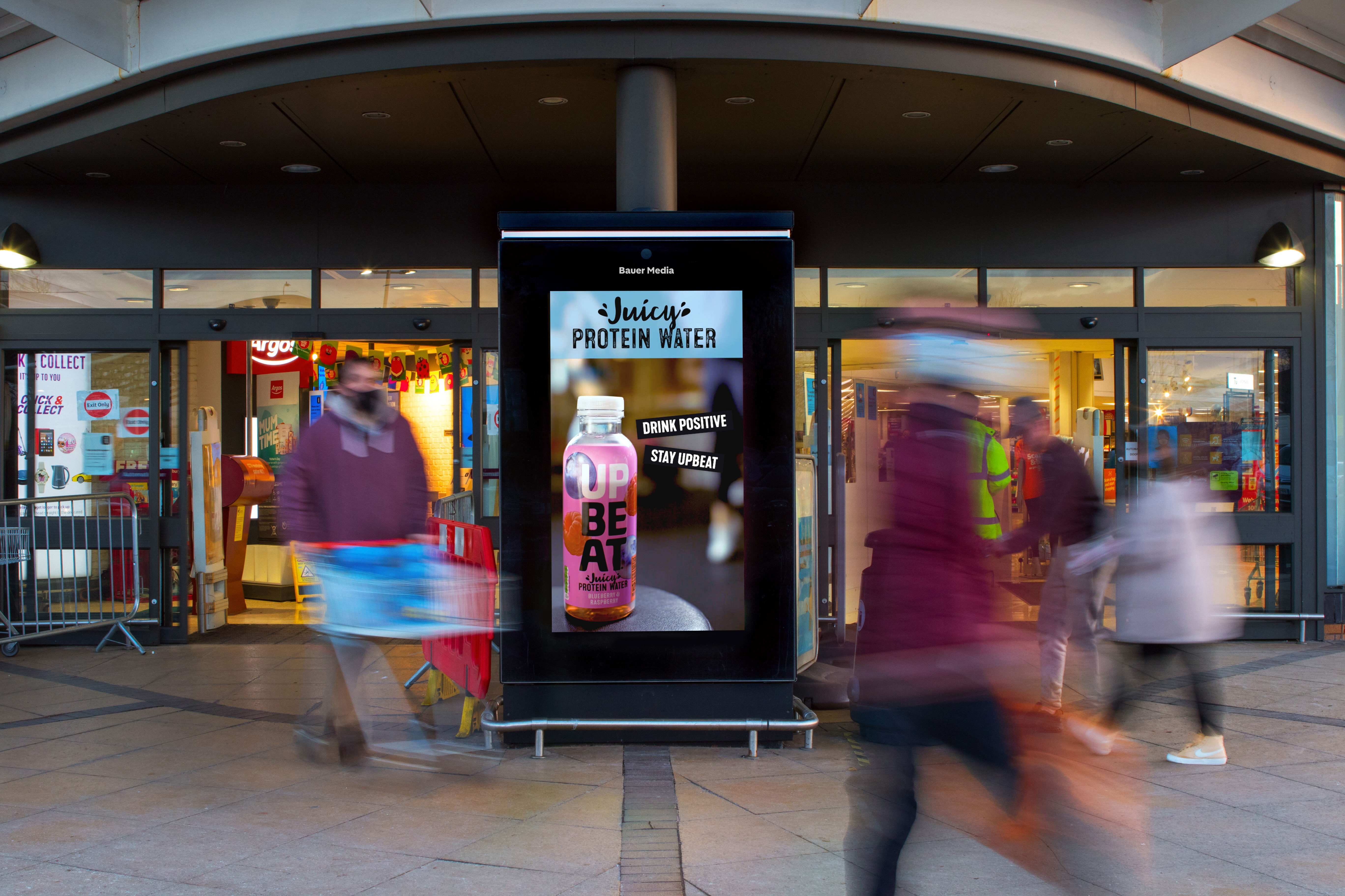 Digital advertising screen showing Juicy Protein Water bottle with text 'Drink Positive Stay Upbeat' outside a store entrance with blurred people walking by.
