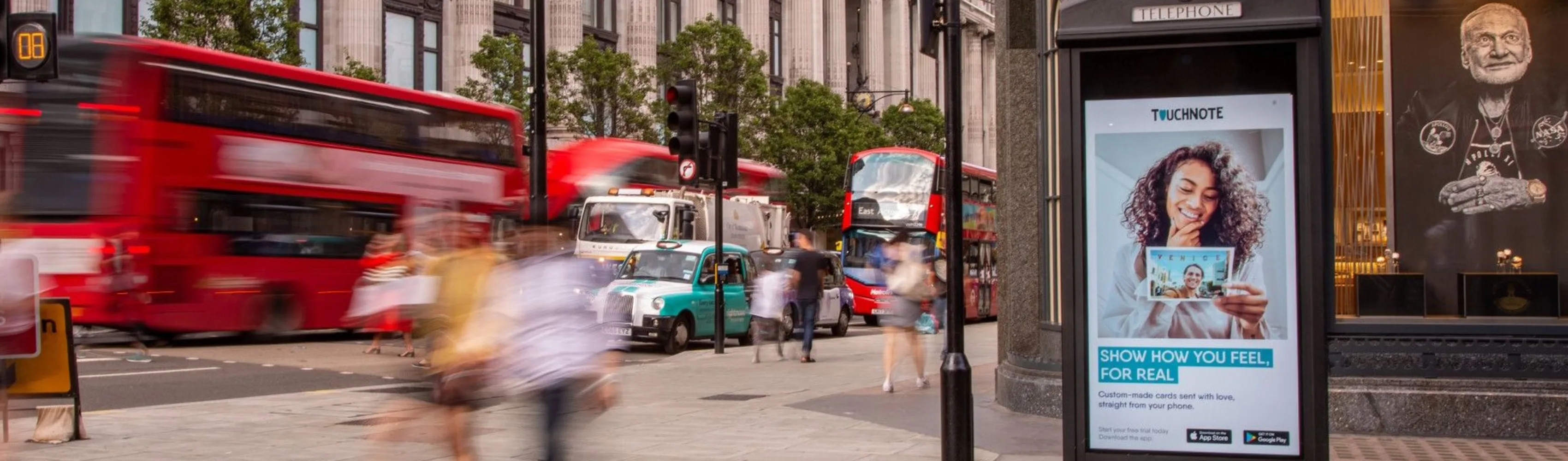 Busy city street with blurred people walking, red double-decker buses, a green taxi, and a digital advertisement showing a smiling woman holding a photo card.