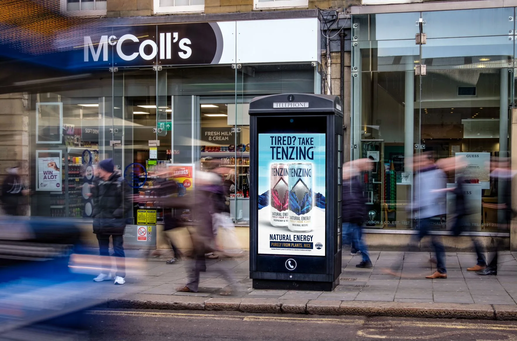 Street scene with blurred pedestrians passing a black telephone booth displaying a Tenzing natural energy drink advertisement in front of a McColl's store.