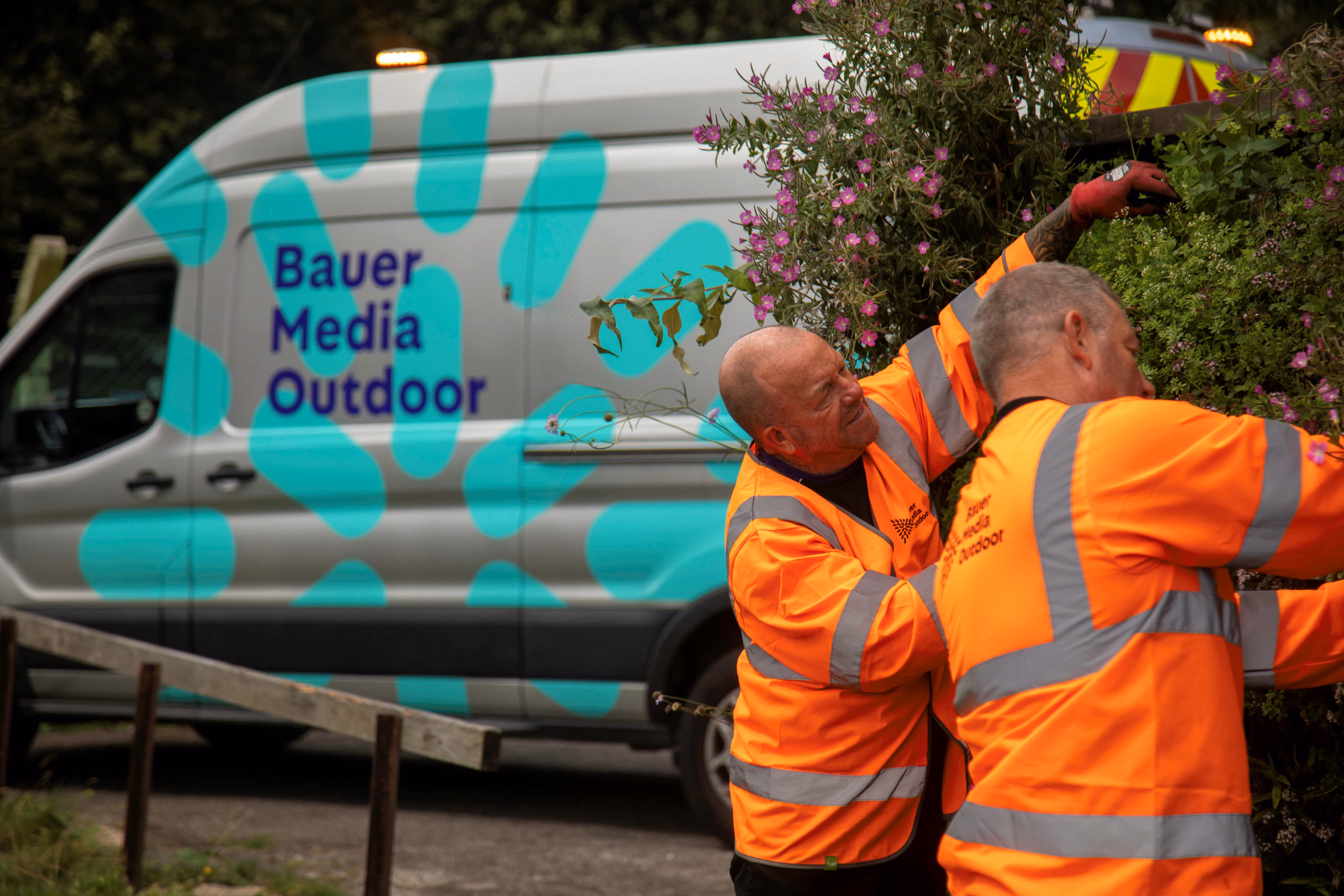 Two workers in bright orange reflective jackets tending to a large vertical garden with purple flowers, next to a van with Bauer Media Outdoor branding.
