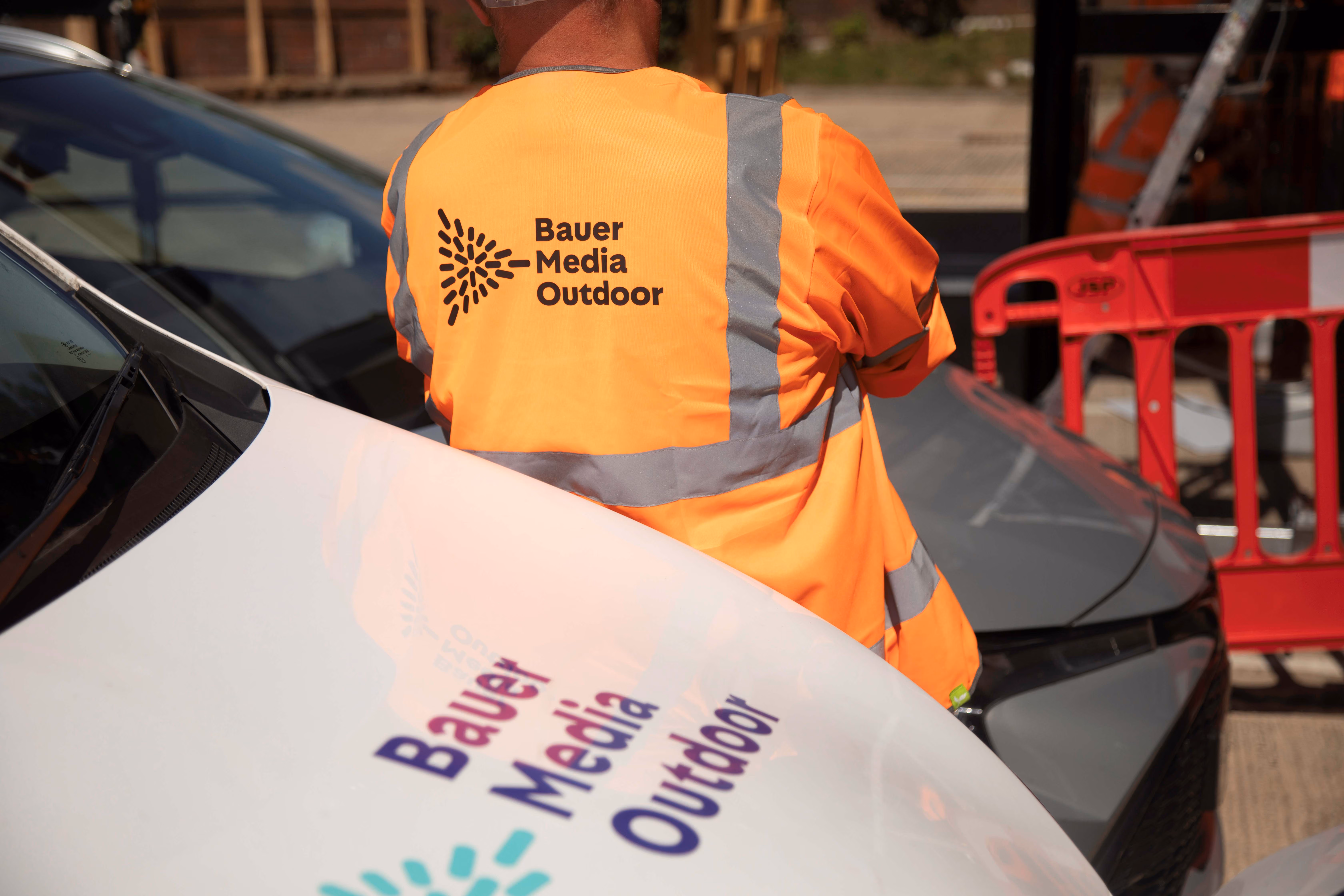 Person wearing an orange Bauer Media Outdoor high-visibility jacket next to a white vehicle with Bauer Media Outdoor logo.