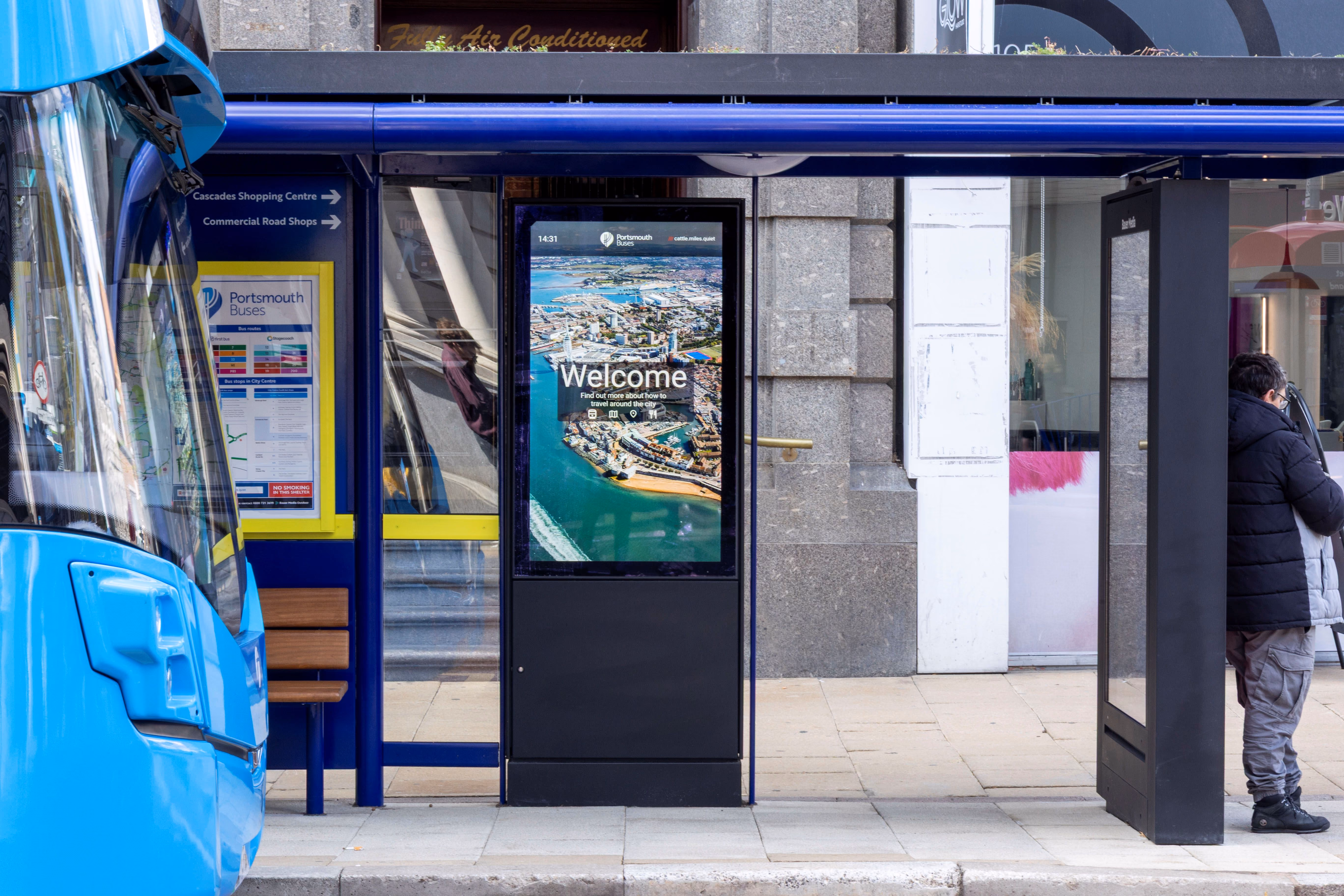 Blue Portsmouth bus parked at a bus stop with a digital welcome sign displaying an aerial view of the city.