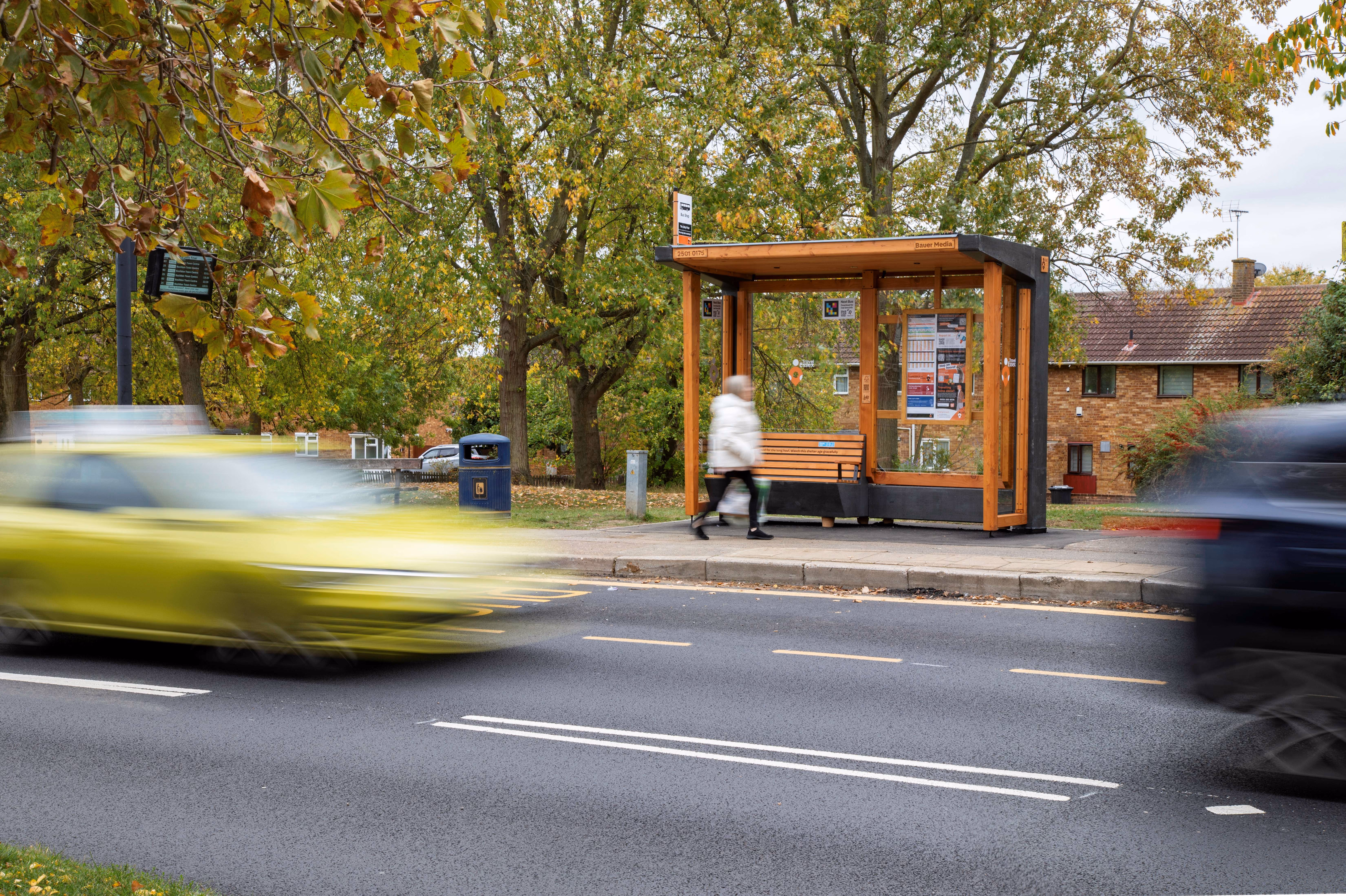 Person walking past a wooden bus shelter on a street with speeding cars and trees in the background.