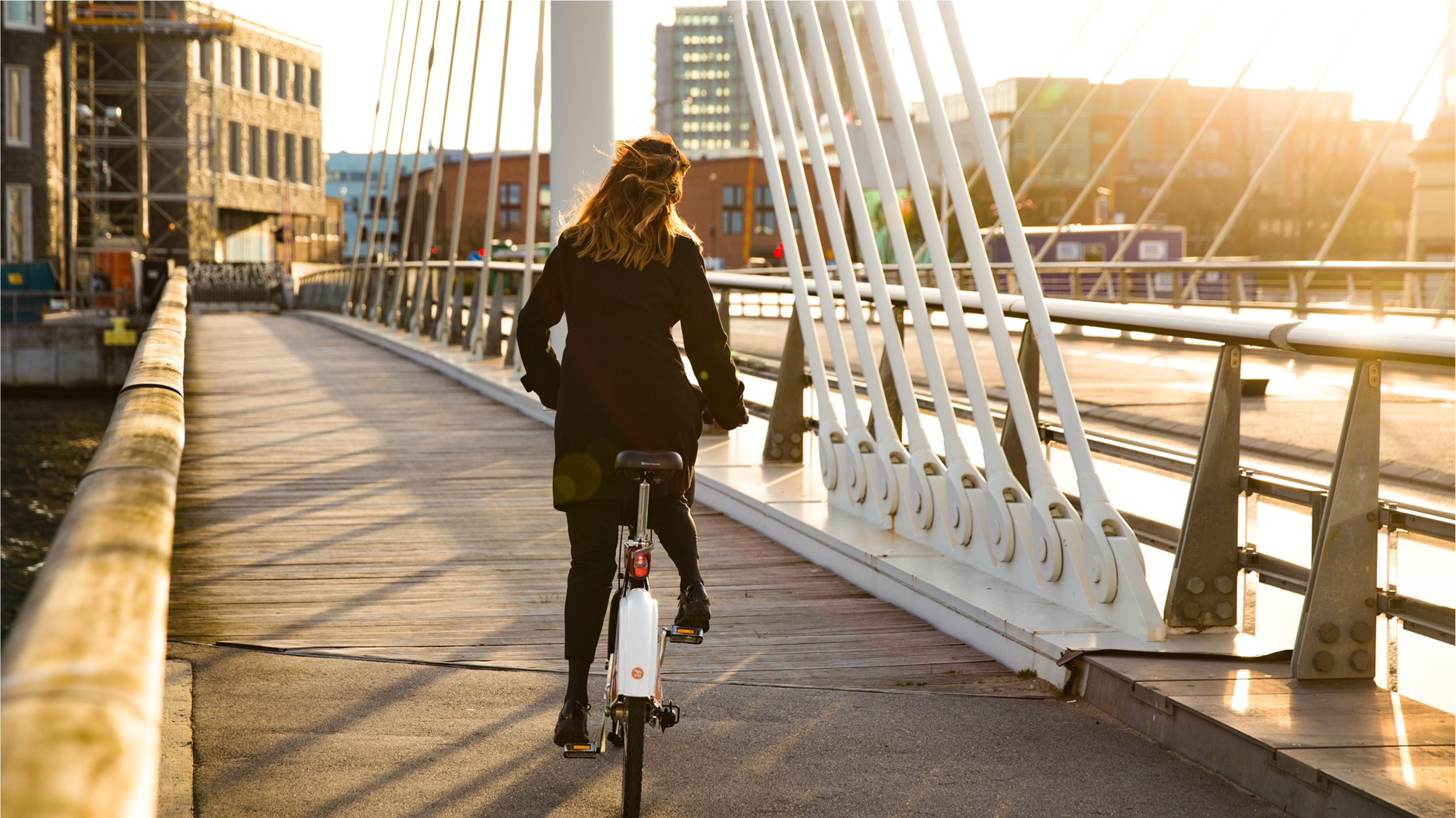 Person wearing a black coat riding a white bicycle on a modern cable-stayed bridge during sunset.