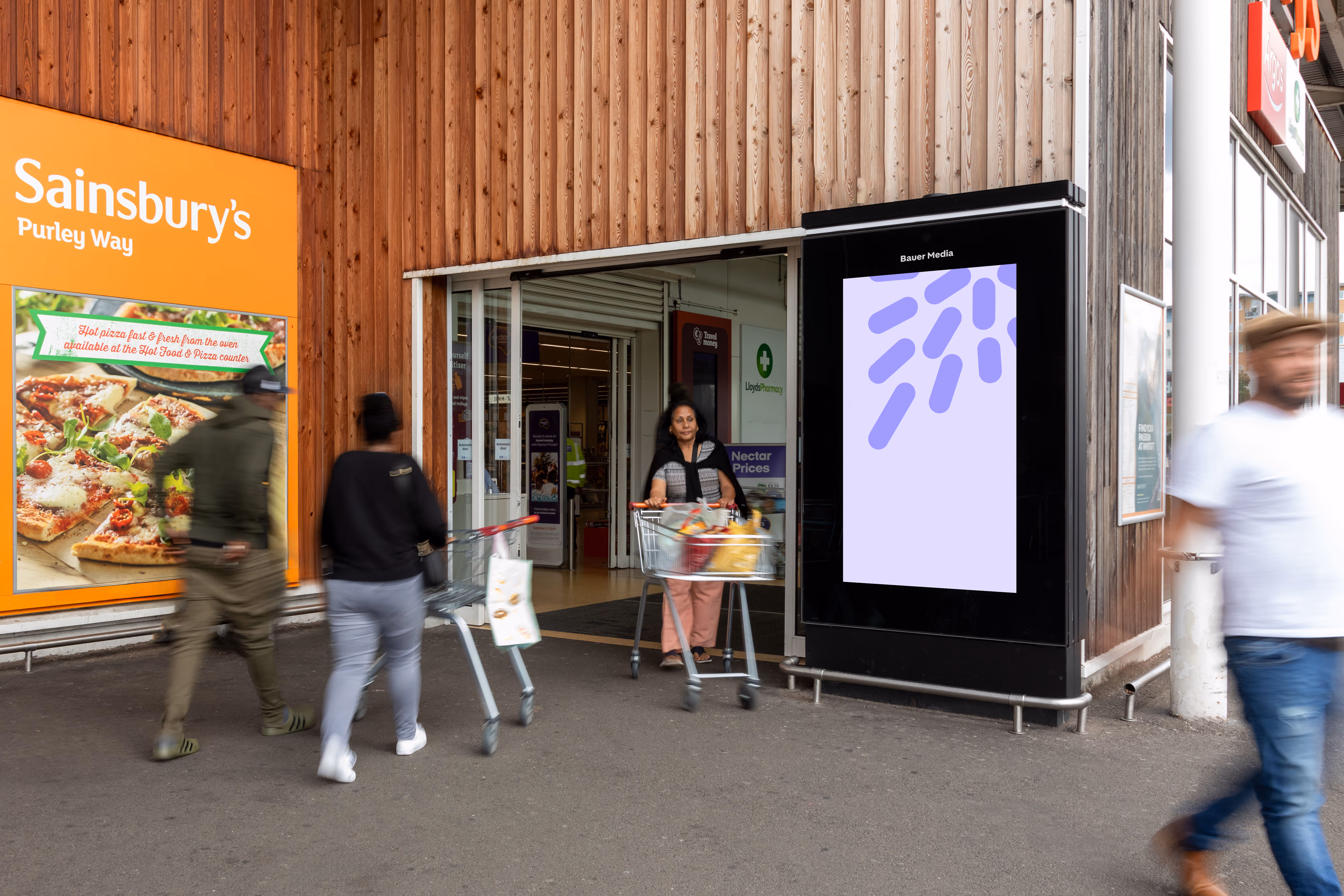 Entrance of a Sainsbury's store with people walking and pushing shopping carts, and a digital advertising screen on the right side.