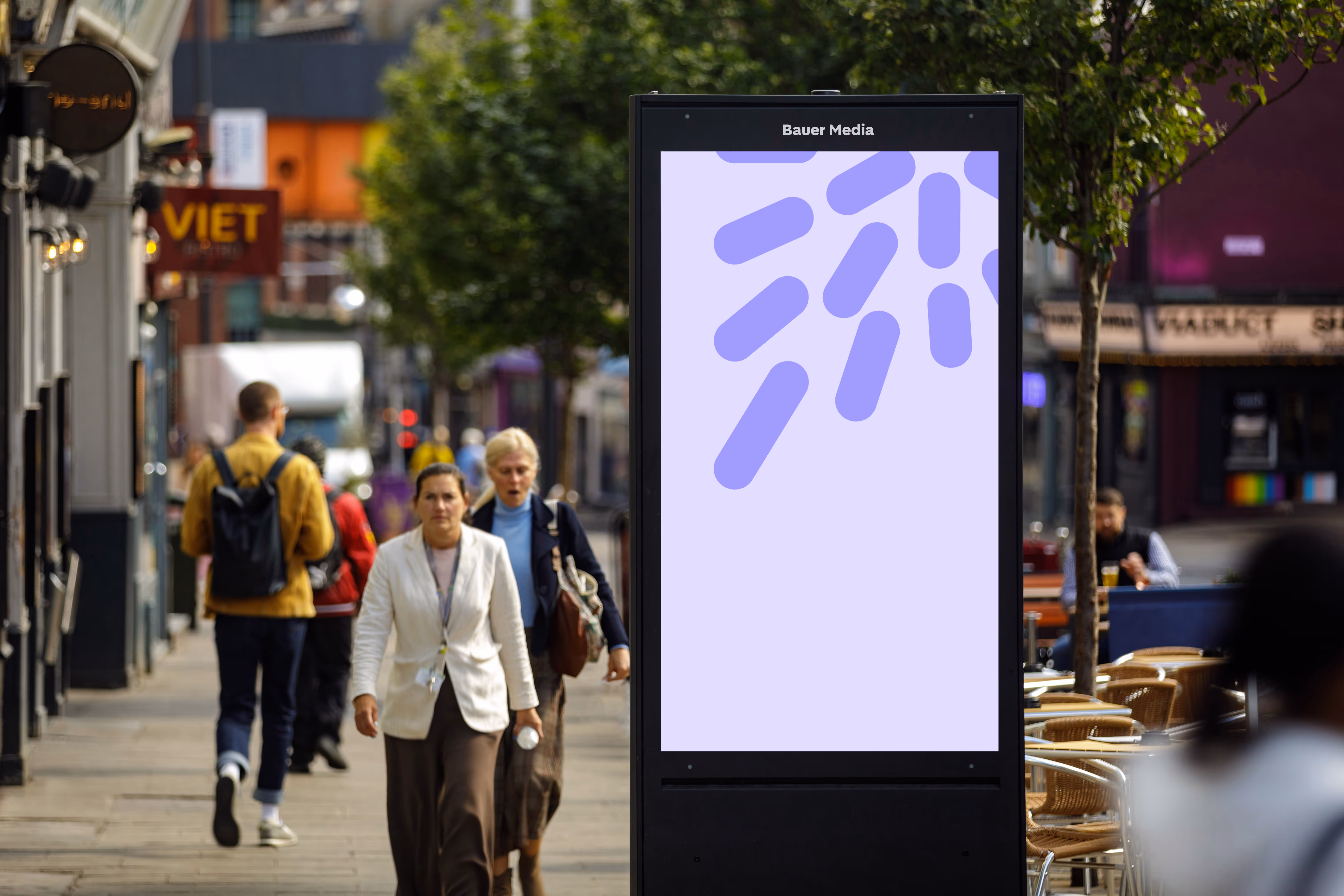 Street scene with people walking on sidewalk beside a digital advertisement panel displaying abstract purple shapes on a light purple background.