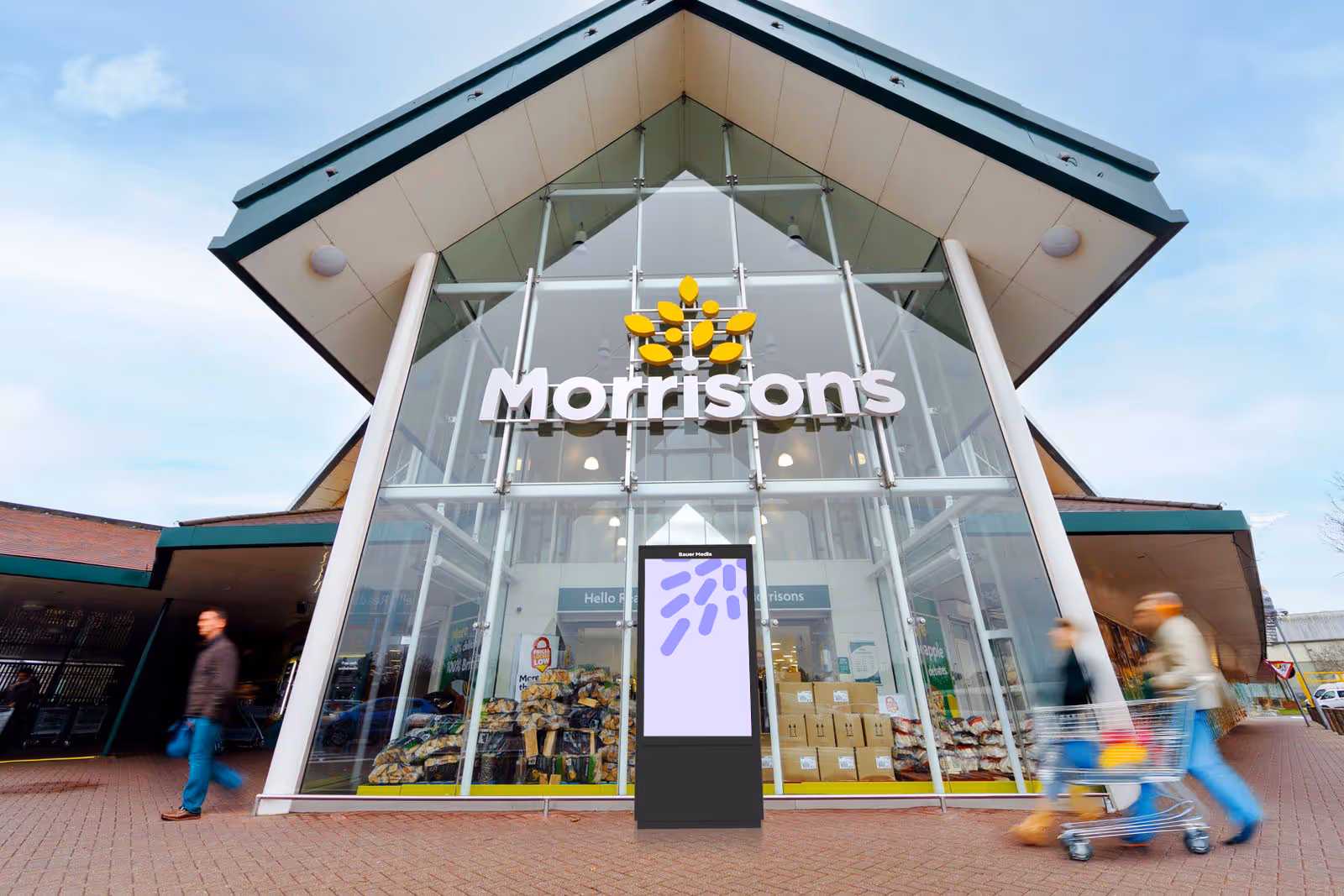 Front entrance of a Morrisons supermarket with glass facade and a digital signboard, with people walking by and pushing a shopping cart.
