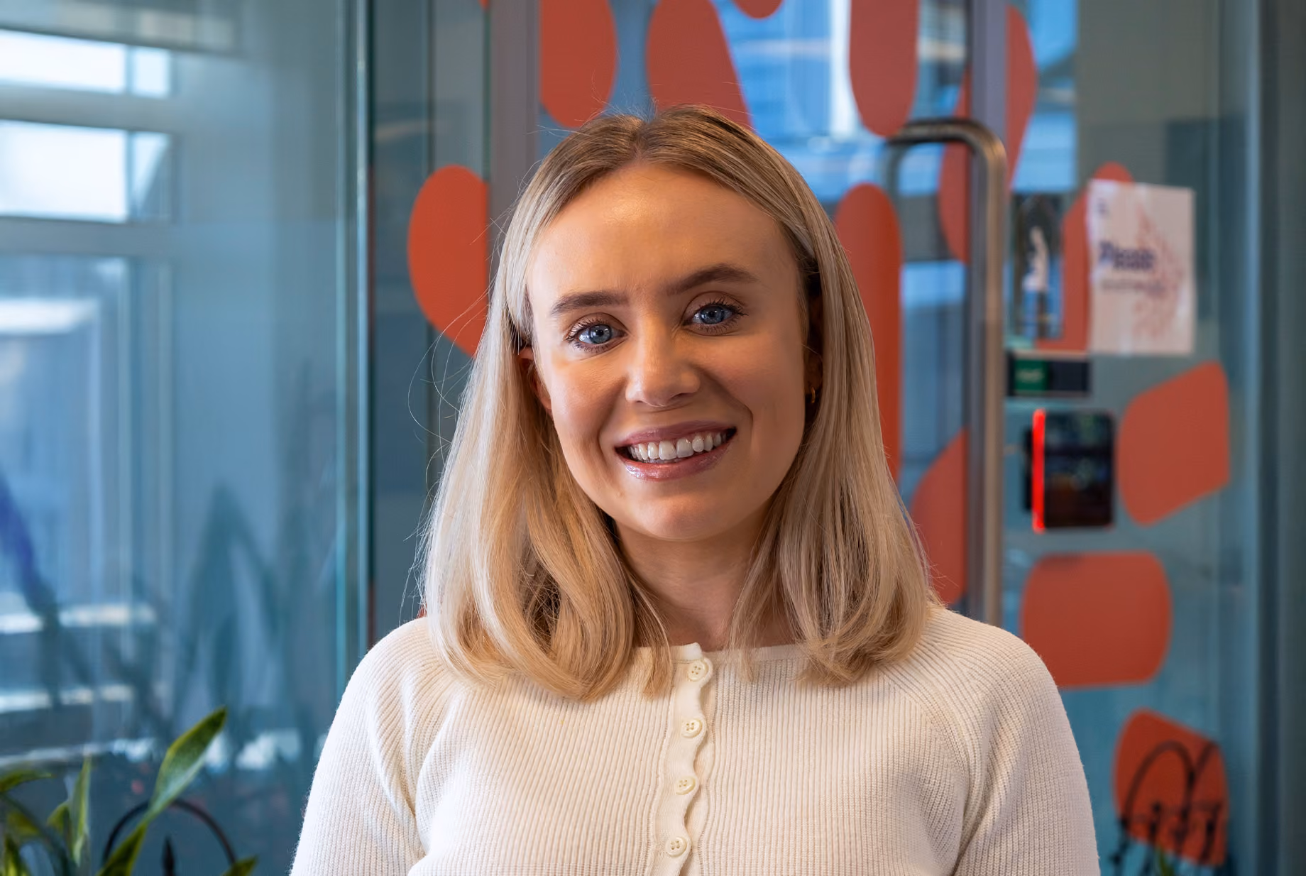 Smiling woman with blonde hair wearing a white buttoned sweater in an office with glass walls.