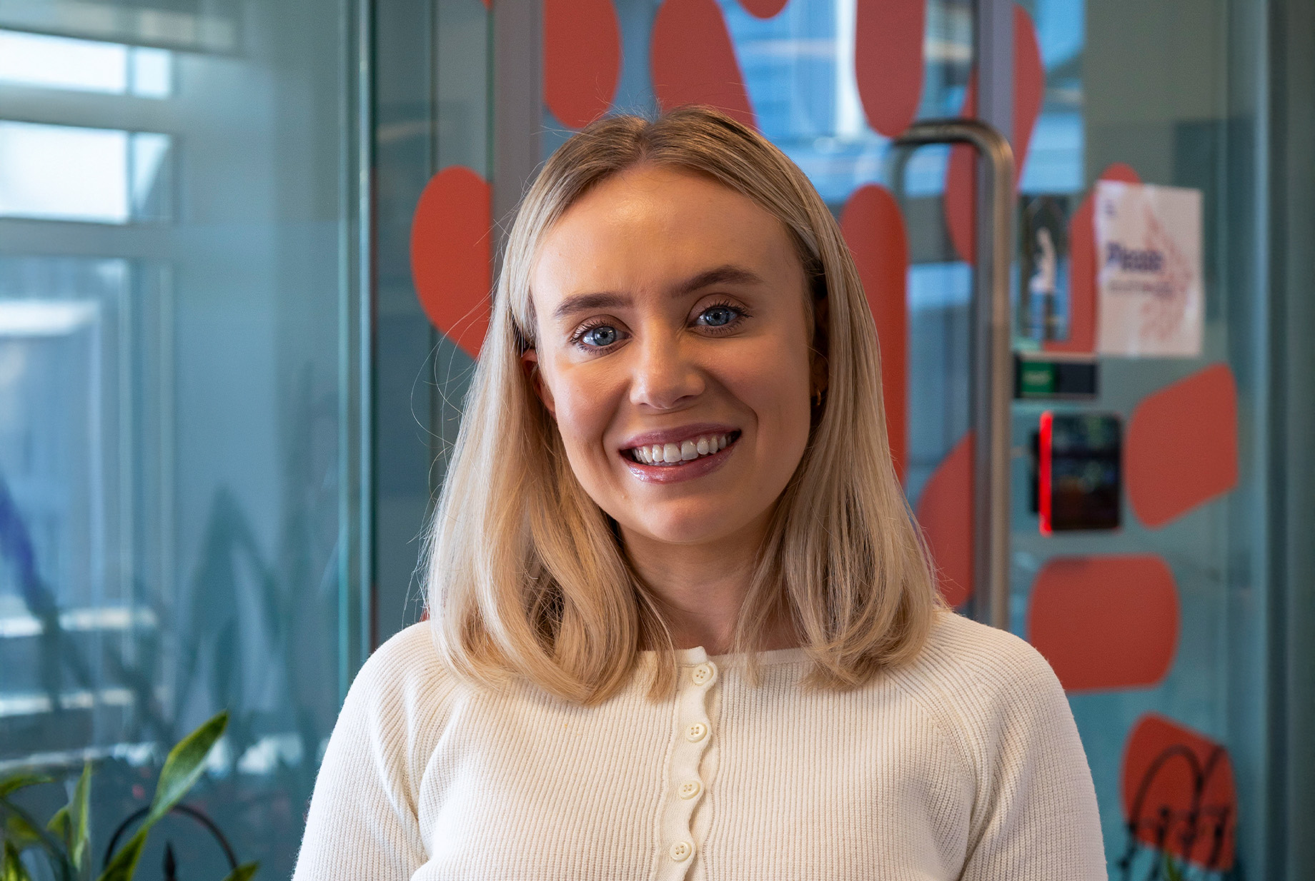 Smiling woman with blonde hair wearing a white buttoned sweater in an office with glass walls.