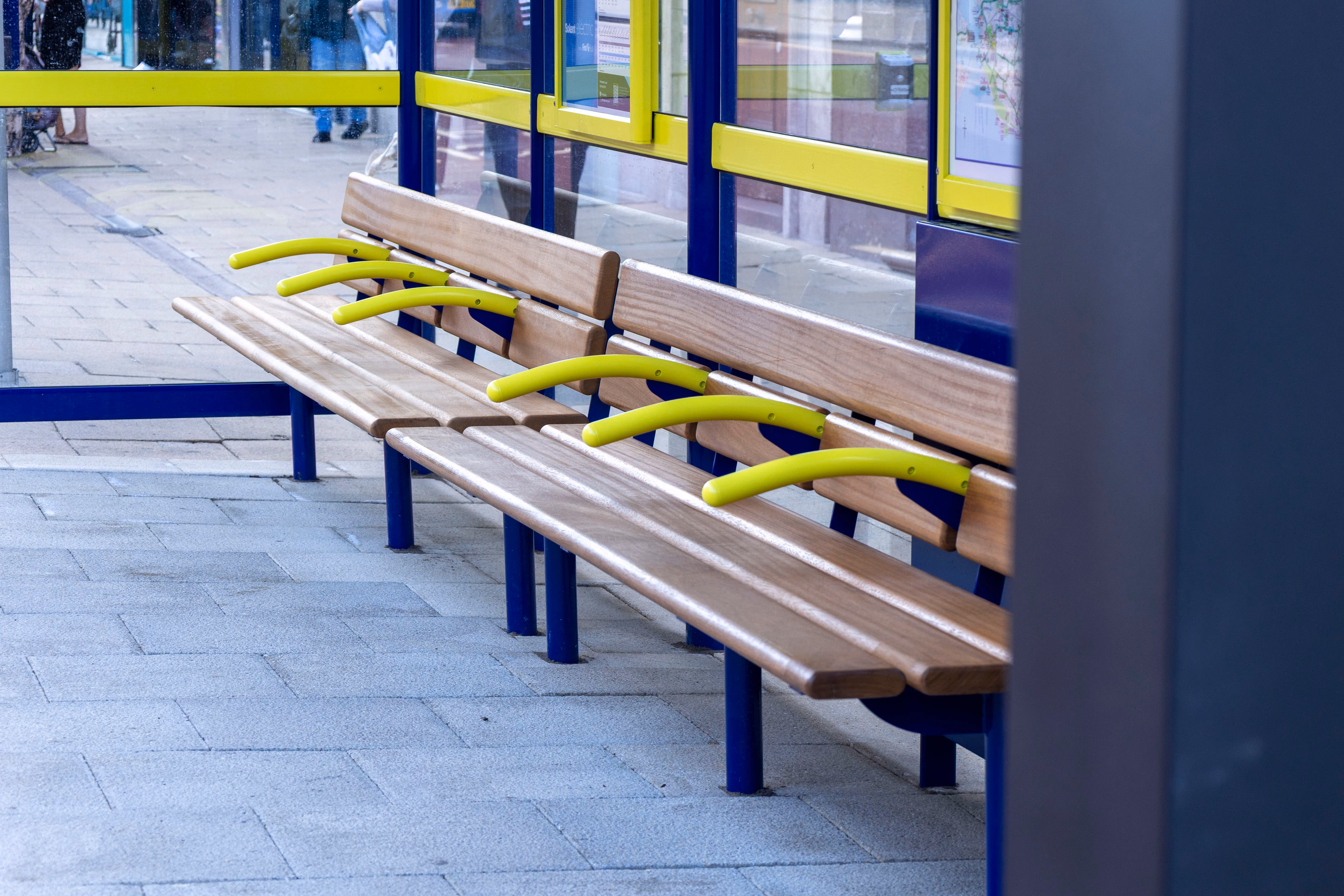 Wooden bench with blue legs and yellow armrests at a bus stop with glass walls and maps.
