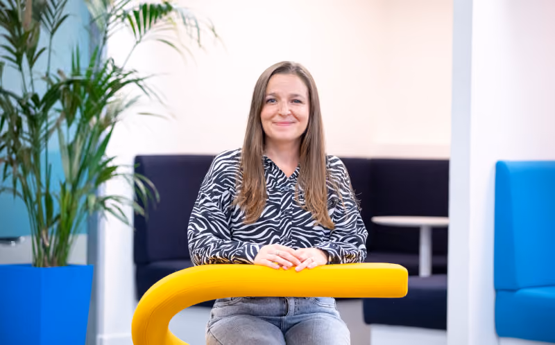 Smiling woman with long hair sitting at a table in a bright room with plants and modern furniture.