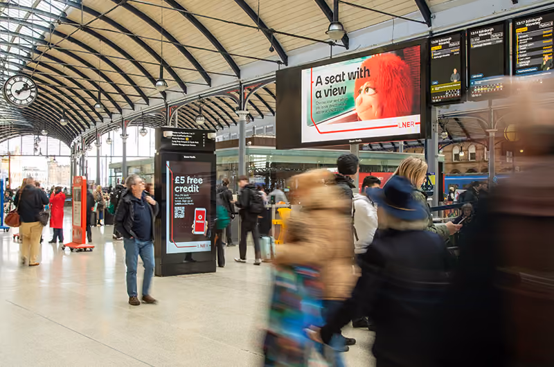 Busy train station concourse with people walking and digital ads, including one for LNER featuring a red-haired animated character.