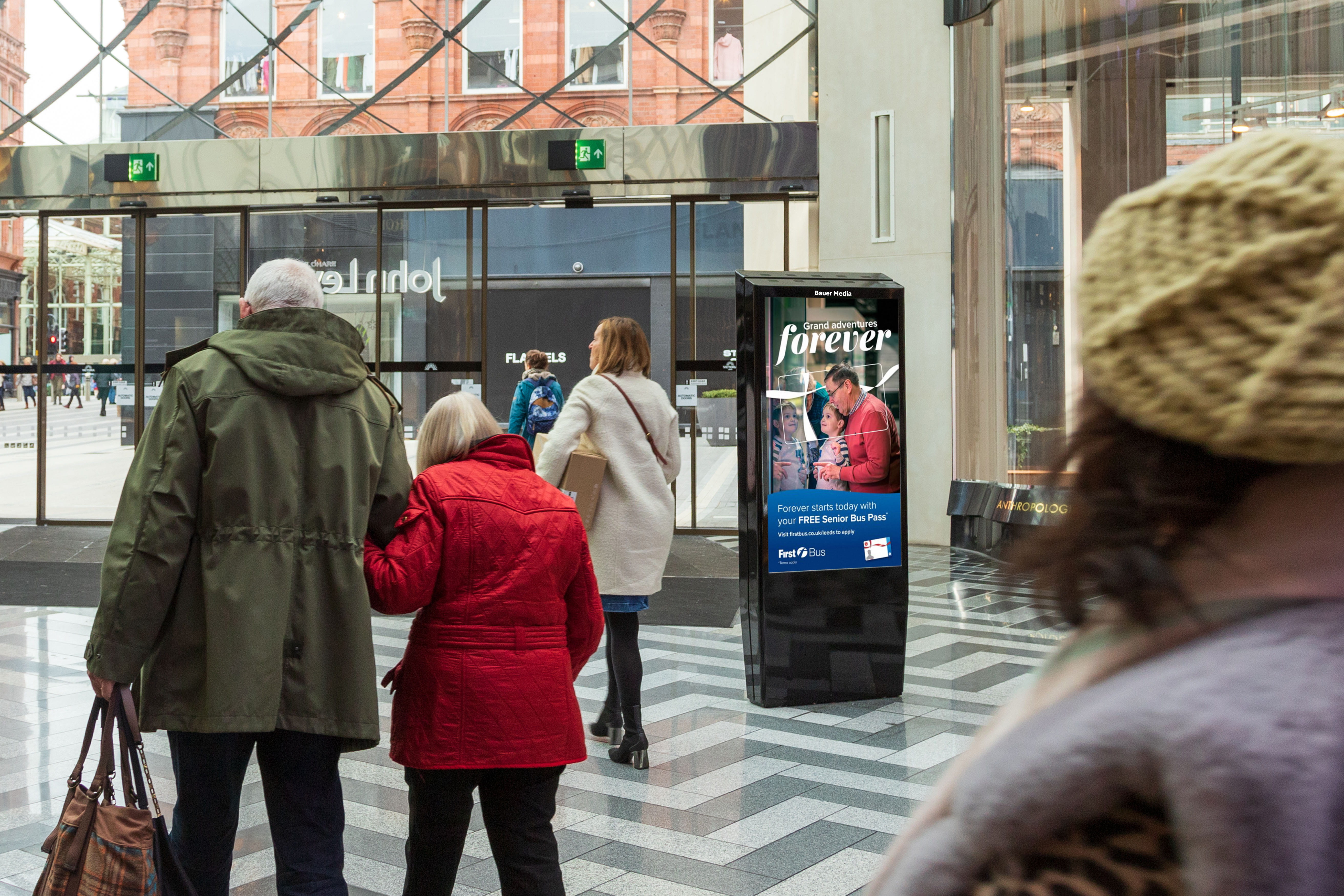 An elderly couple walking arm in arm inside a modern building near a digital advertisement for a free senior bus pass.