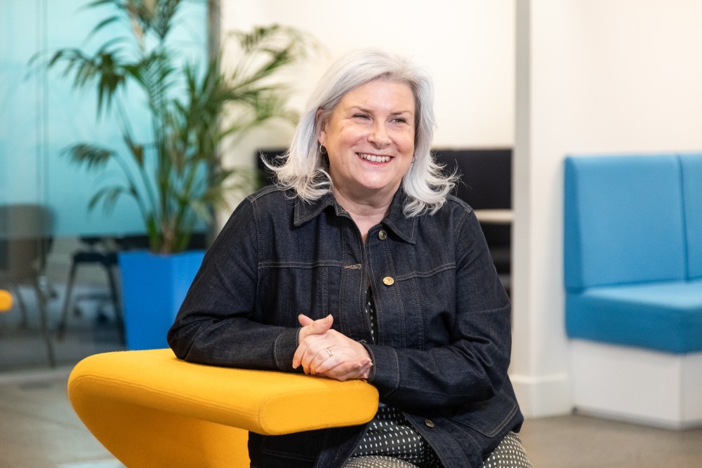 Smiling woman with gray hair wearing a dark denim jacket, sitting on a yellow chair in a modern office space with plants and blue seating in the background.
