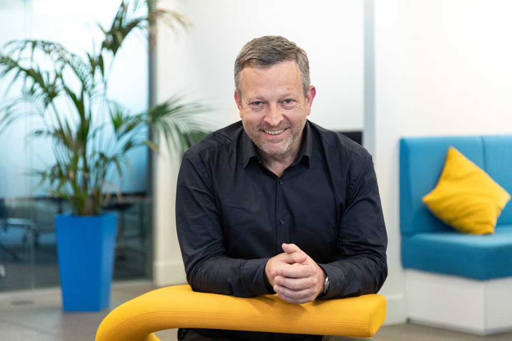 Man in a black shirt smiling while leaning on a yellow chair in a modern office with blue furniture and a potted plant in the background.