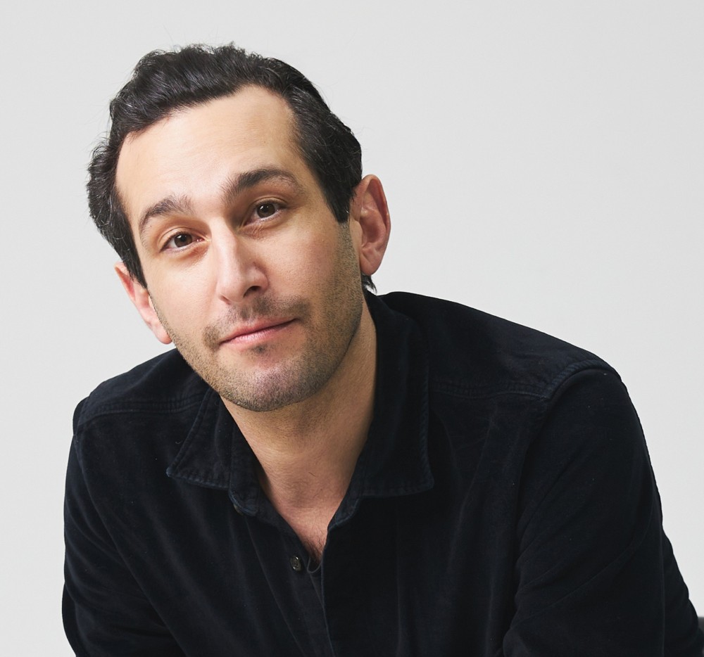 Man with dark hair and light beard wearing a black collared shirt against a plain light background.