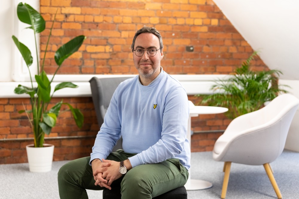 Man wearing glasses, a light blue sweatshirt, and green pants sitting in a modern office space with brick walls and potted plants.