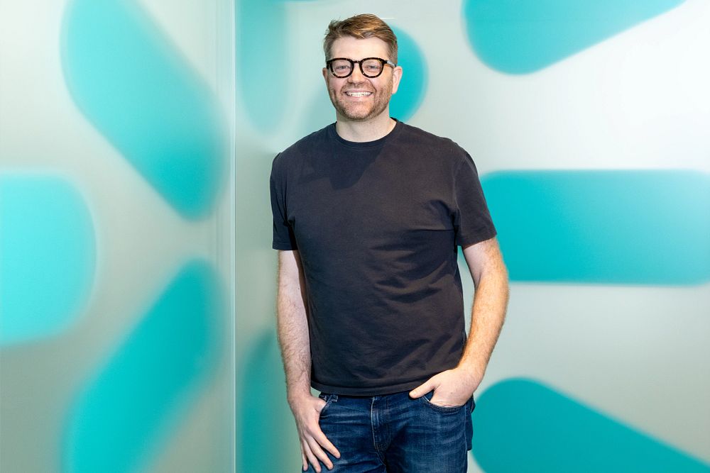 Smiling man with glasses wearing a black t-shirt and jeans standing against a light-colored wall with blue abstract shapes.