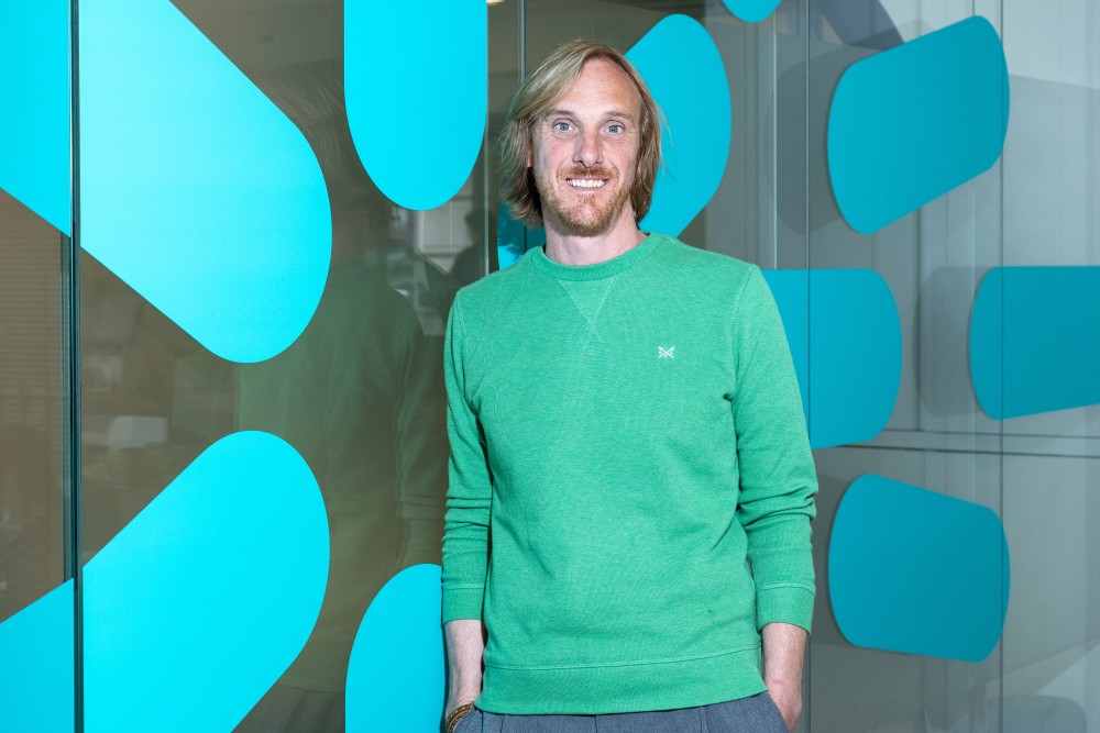 Smiling man with light brown hair and beard wearing a green sweater, standing in front of a glass wall with blue abstract shapes.